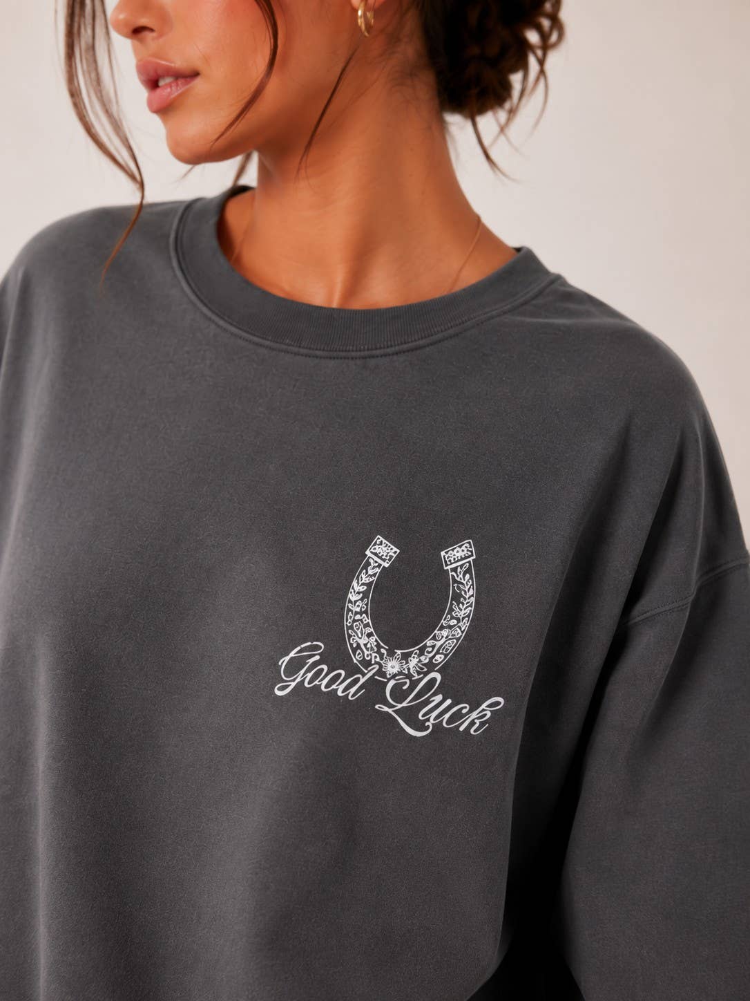 A close-up of a woman wearing a dark gray crewneck sweatshirt with a white embroidered horseshoe and 'Good Luck' text on the chest.