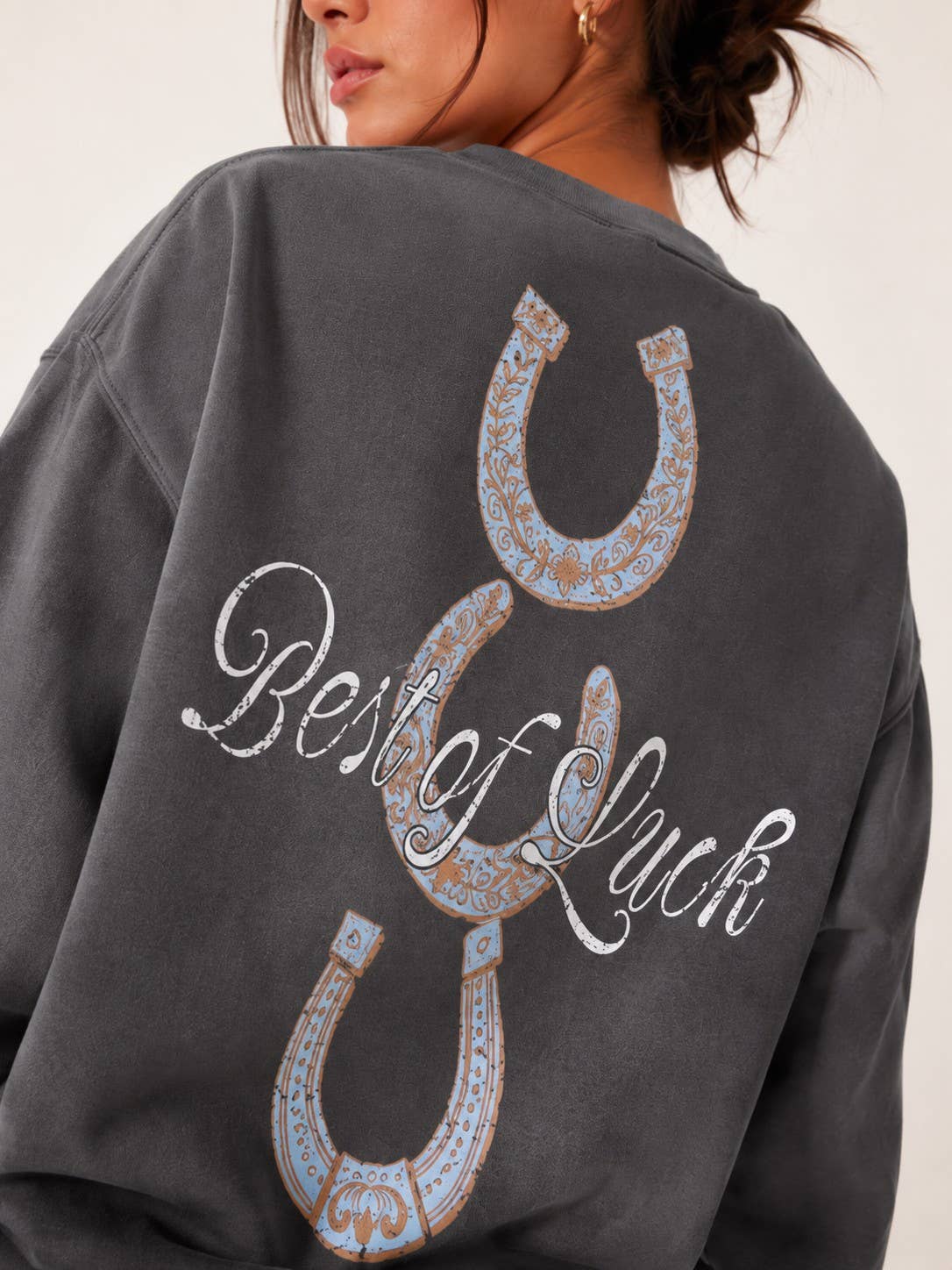 A close-up of a person wearing a dark gray sweatshirt, showing the back of the garment with a large graphic of three horseshoes and the words 'Best of Luck' in white script.