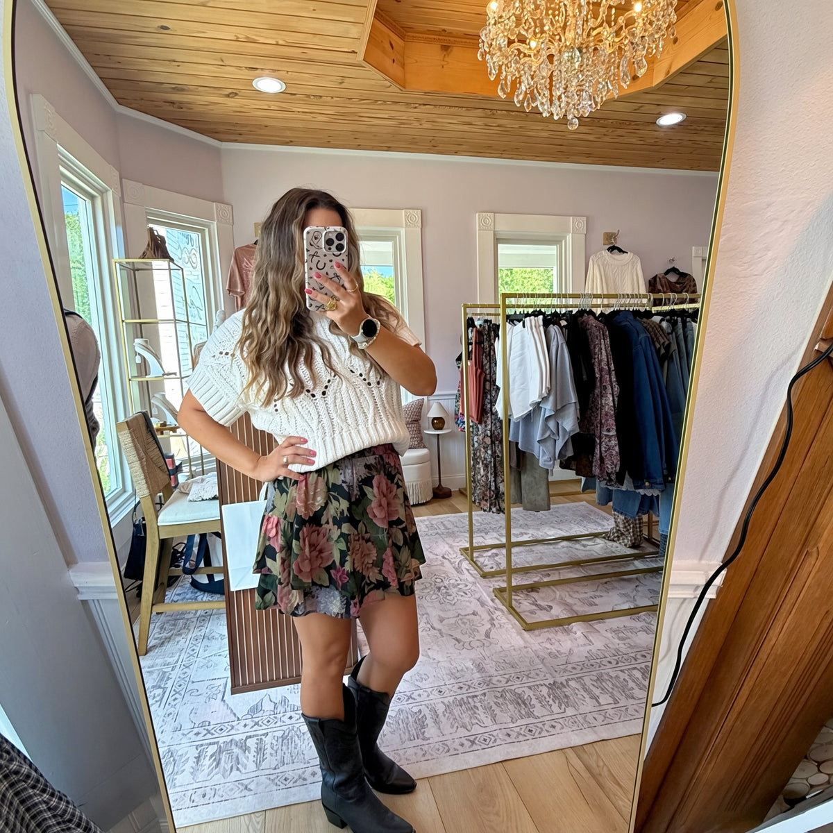 A woman takes a mirror selfie in a boutique clothing store, wearing a white knit short-sleeve top with a floral print mini skirt and black knee-high boots. The boutique features a wood ceiling, crystal chandelier, and clothing racks in the background.