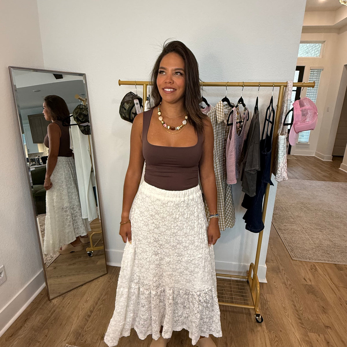 Woman in a brown top and white lace skirt standing in a room with a full-length mirror and clothing rack.