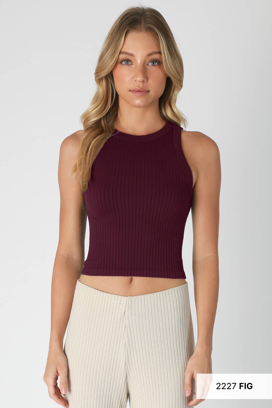 A woman modeling a sleeveless ribbed knit crop top in deep burgundy, paired with cream ribbed pants, against a light gray studio background.