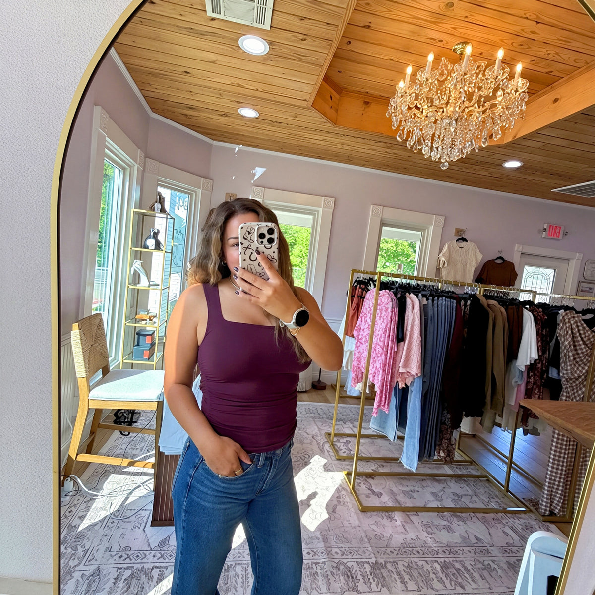 A woman takes a mirror selfie in a boutique, wearing a sleeveless plum purple tank top and blue jeans. The boutique features a wooden ceiling, a crystal chandelier, clothing racks with various garments, and a patterned rug.