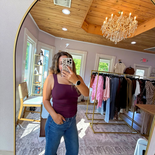 A woman takes a mirror selfie in a boutique, wearing a sleeveless plum purple tank top and blue jeans. The boutique features a wooden ceiling, a crystal chandelier, clothing racks with various garments, and a patterned rug.