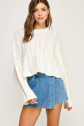 A woman modeling a white cable-knit long-sleeve sweater paired with a blue denim mini skirt, set against a plain light gray studio background.