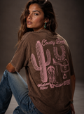 Woman wearing a brown t-shirt with a cactus and boot design on the back, sitting against a gray background.