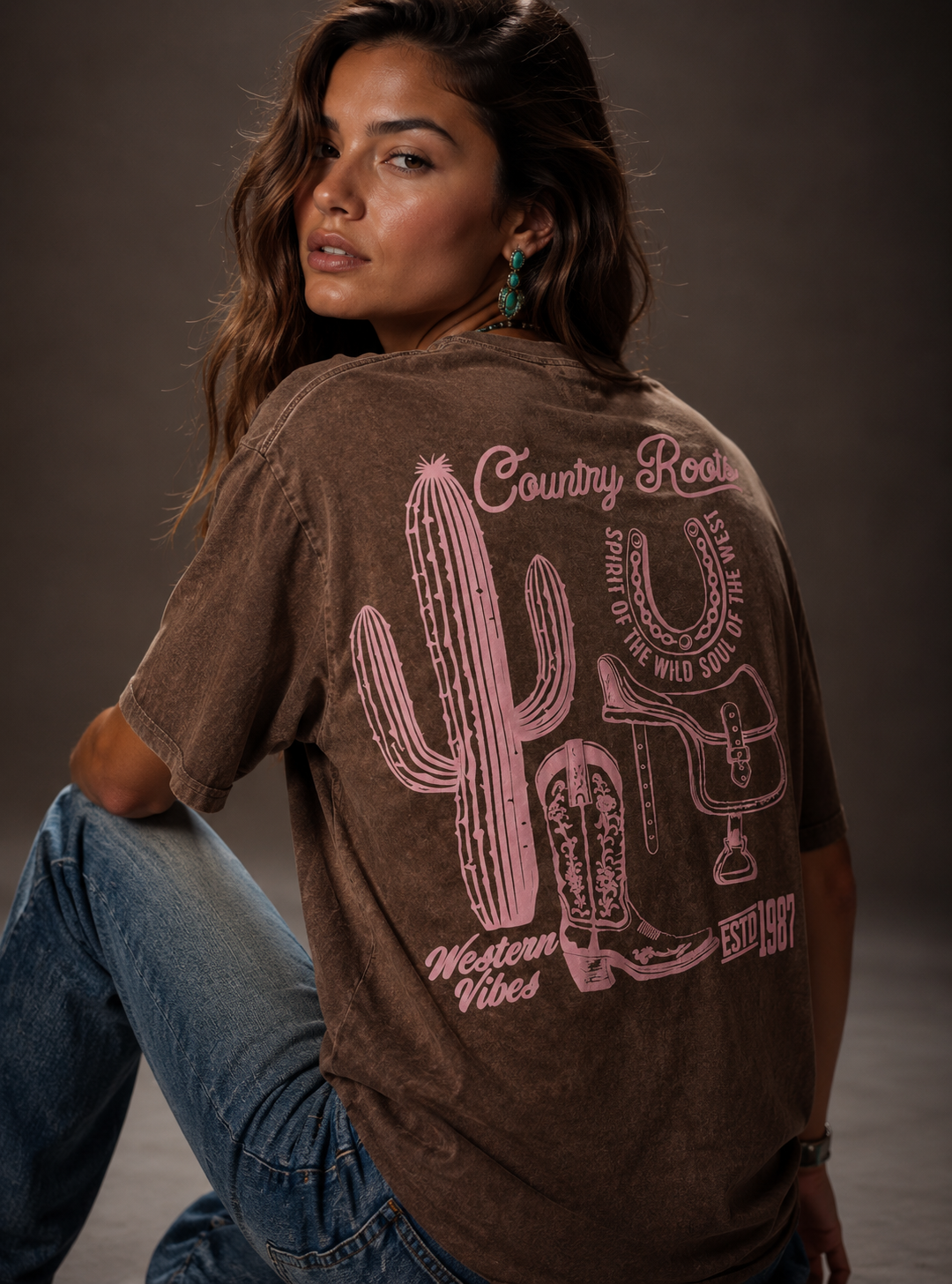 Woman wearing a brown t-shirt with a cactus and boot design on the back, sitting against a gray background.
