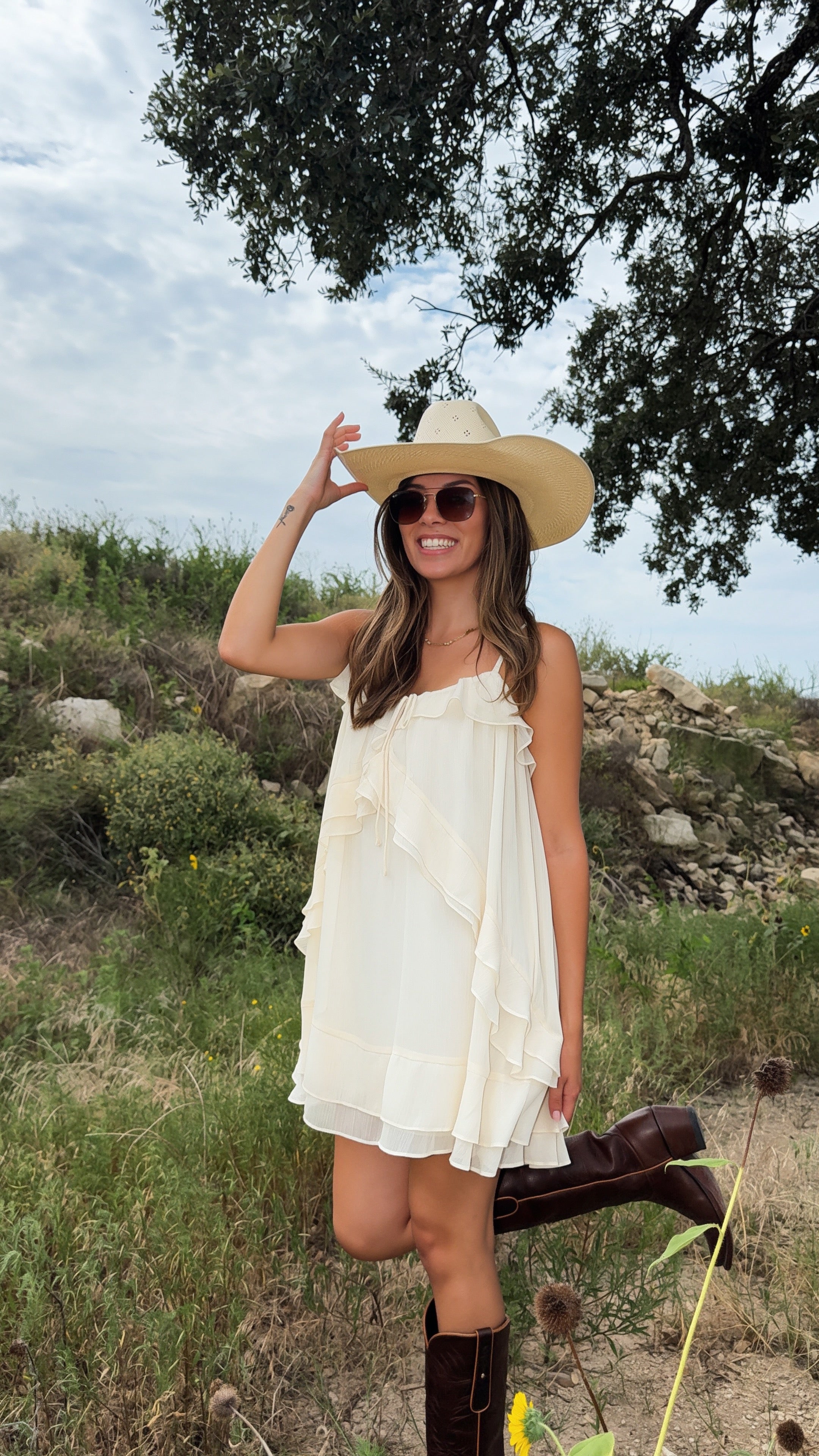 A woman wearing a cream ruffled sleeveless western style fall dress, a wide brim straw cowboy hat, and brown cowgirl boots stands outdoors in a grassy field under a cloudy sky.