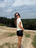 A woman wearing a white textured short-sleeve top and a black faux-leather mini skirt, standing outdoors on a dirt path with a stone wall and trees in the background.