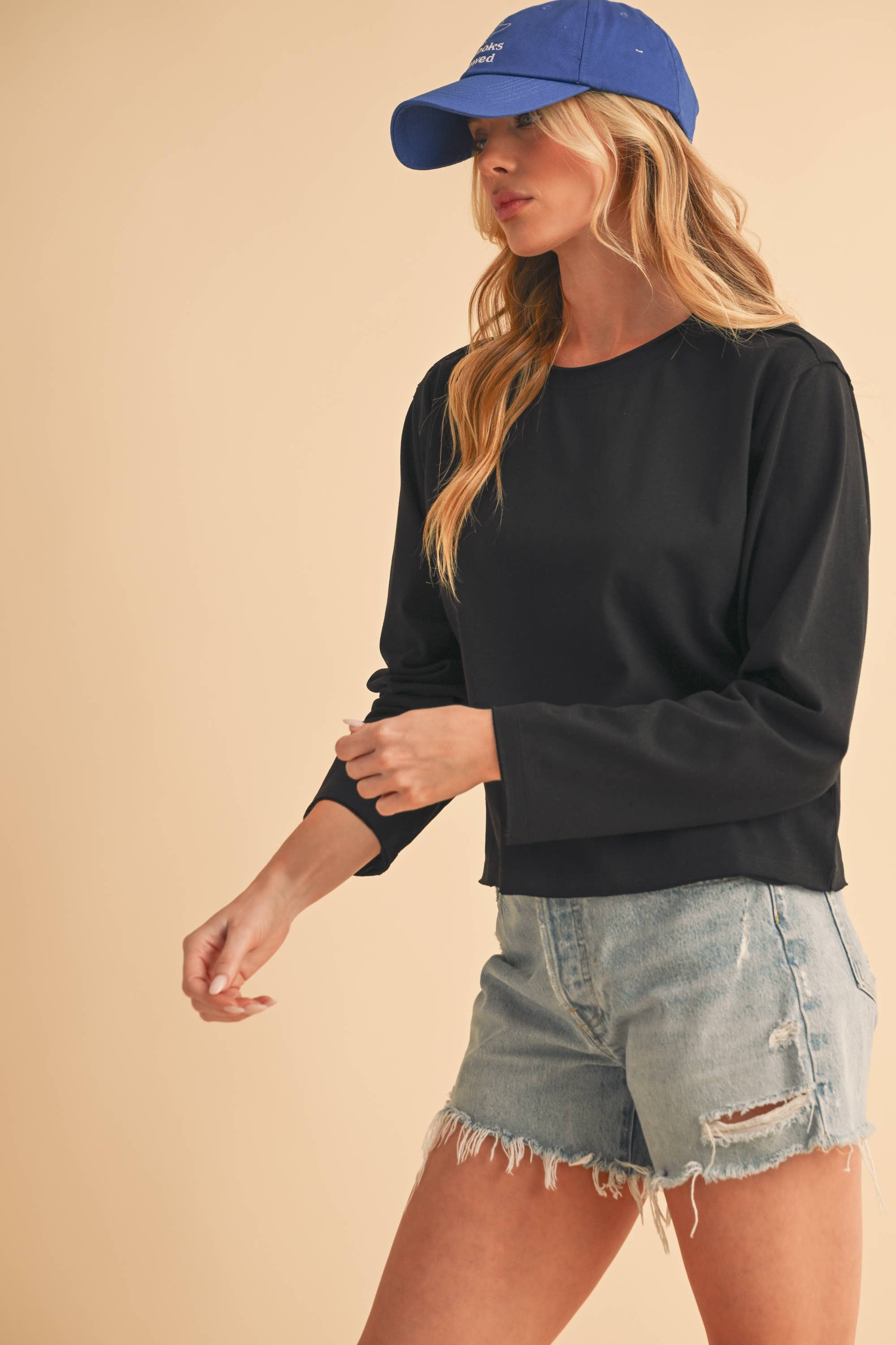 Woman wearing a black long sleeve crop top, blue cap, and denim shorts on a beige background