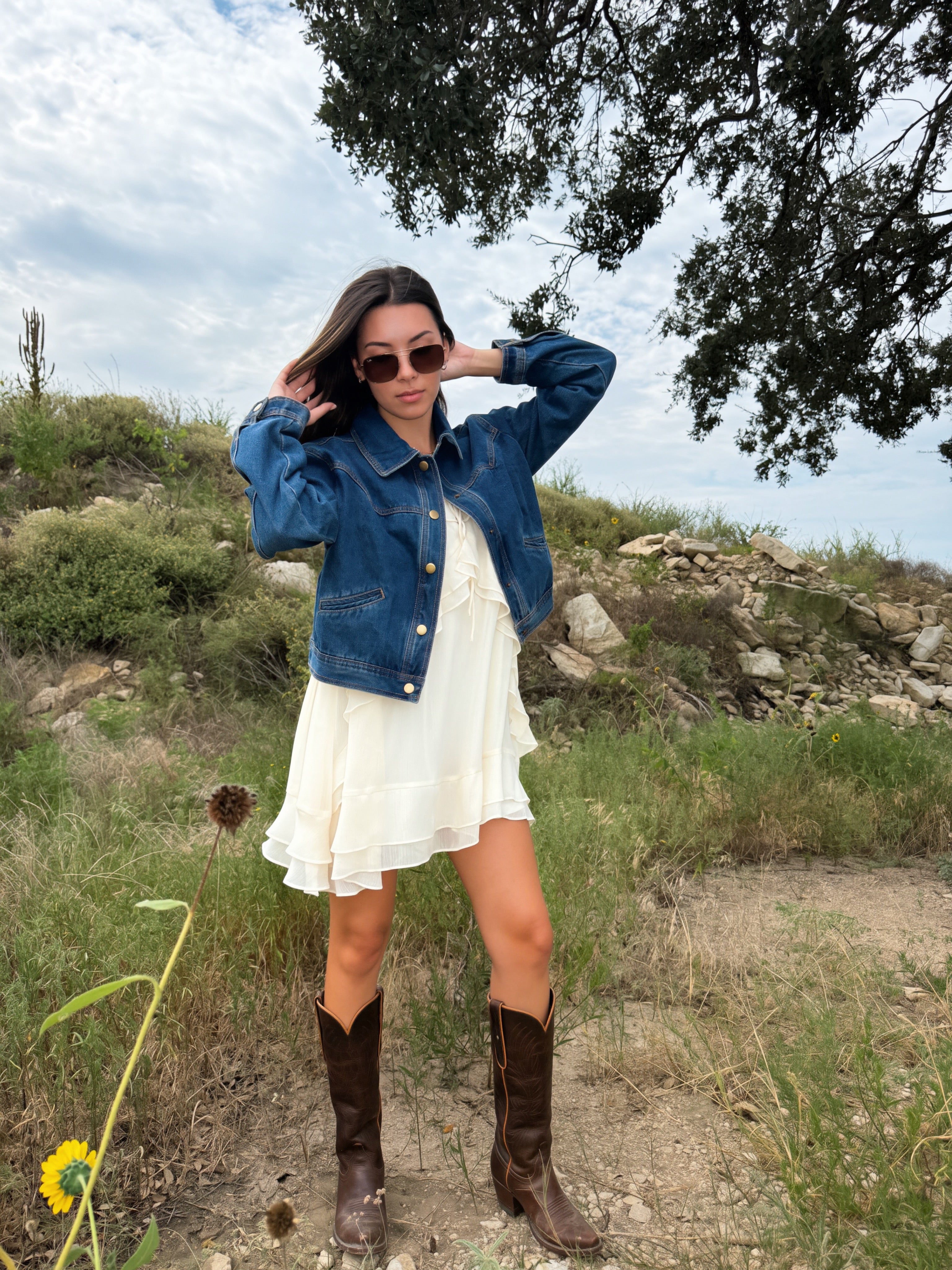 A woman stands outdoors on a grassy hill wearing a blue fall western style denim jacket over a white ruffled dress, with brown cowboy boots and sunglasses.