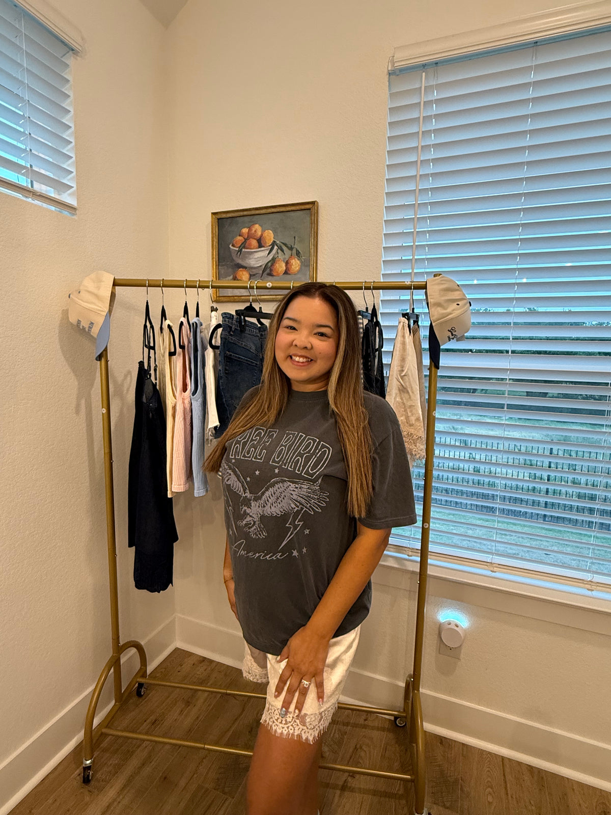 A woman stands in a room wearing a dark gray graphic t-shirt and white lace-trimmed shorts, posing next to a gold clothing rack filled with various garments.