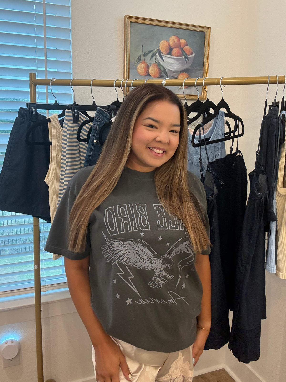 A smiling young woman stands indoors in front of a clothing rack, wearing a dark gray graphic T-shirt with an eagle design and the words 'BIRD' and 'America' visible on the front, paired with light beige pants.