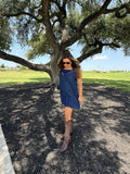 A woman wearing a sleeveless navy blue dress and brown cowboy boots stands outdoors under a large tree on a paved path with grass in the background.