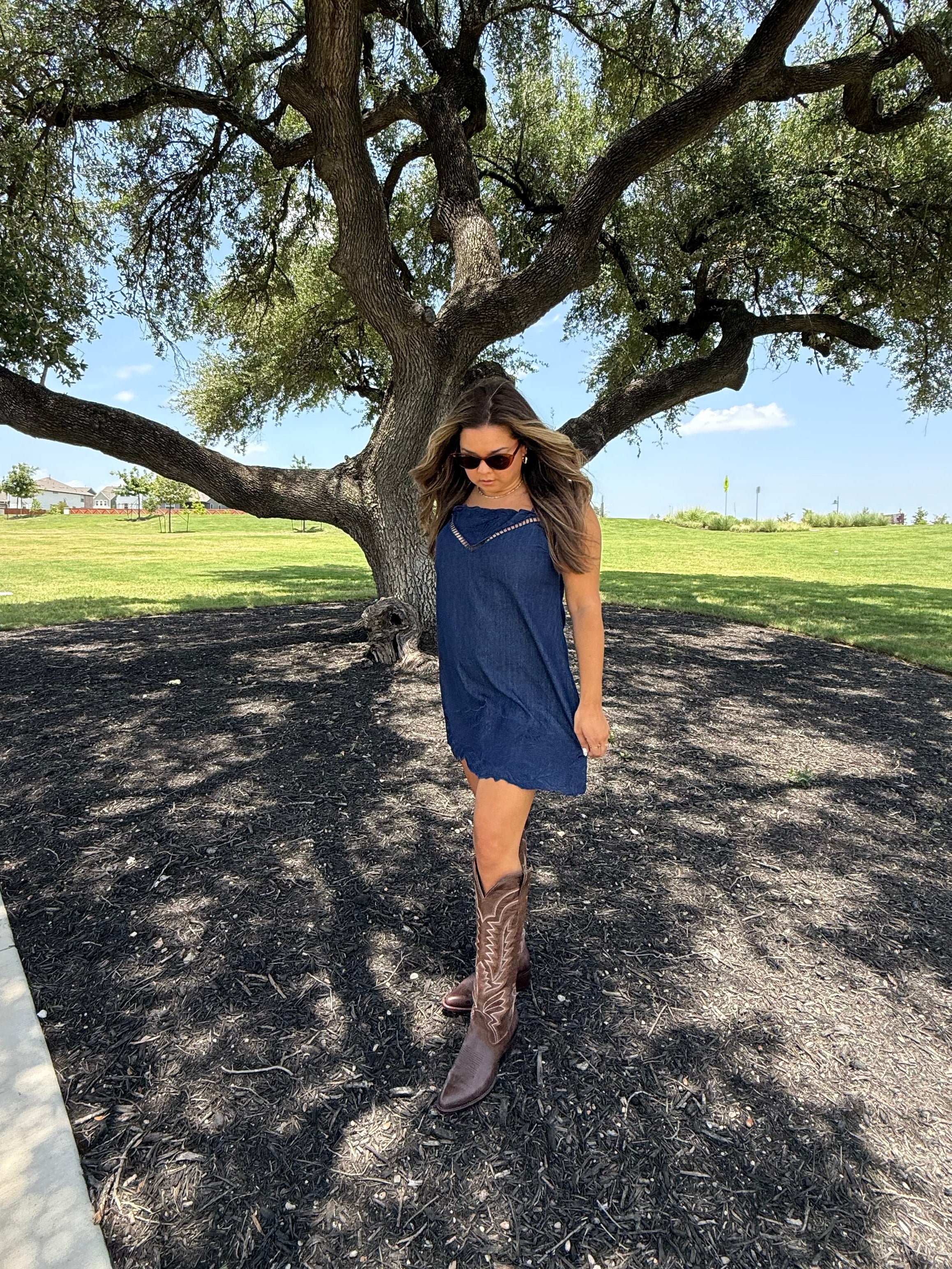 A woman wearing a sleeveless navy blue dress and brown cowboy boots stands outdoors under a large tree on a paved path with grass in the background.