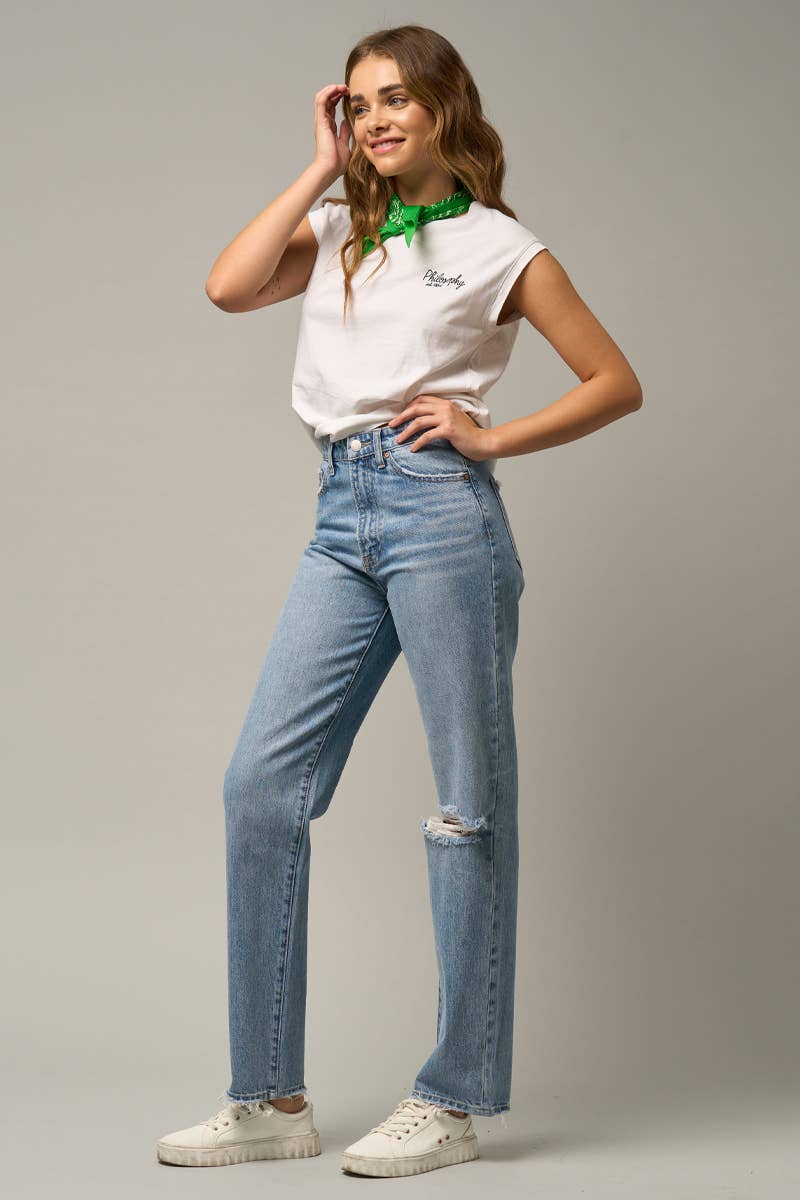 A woman modeling a white short-sleeve t-shirt with a green neck scarf, light-wash distressed blue jeans, and white sneakers against a solid gray studio background.