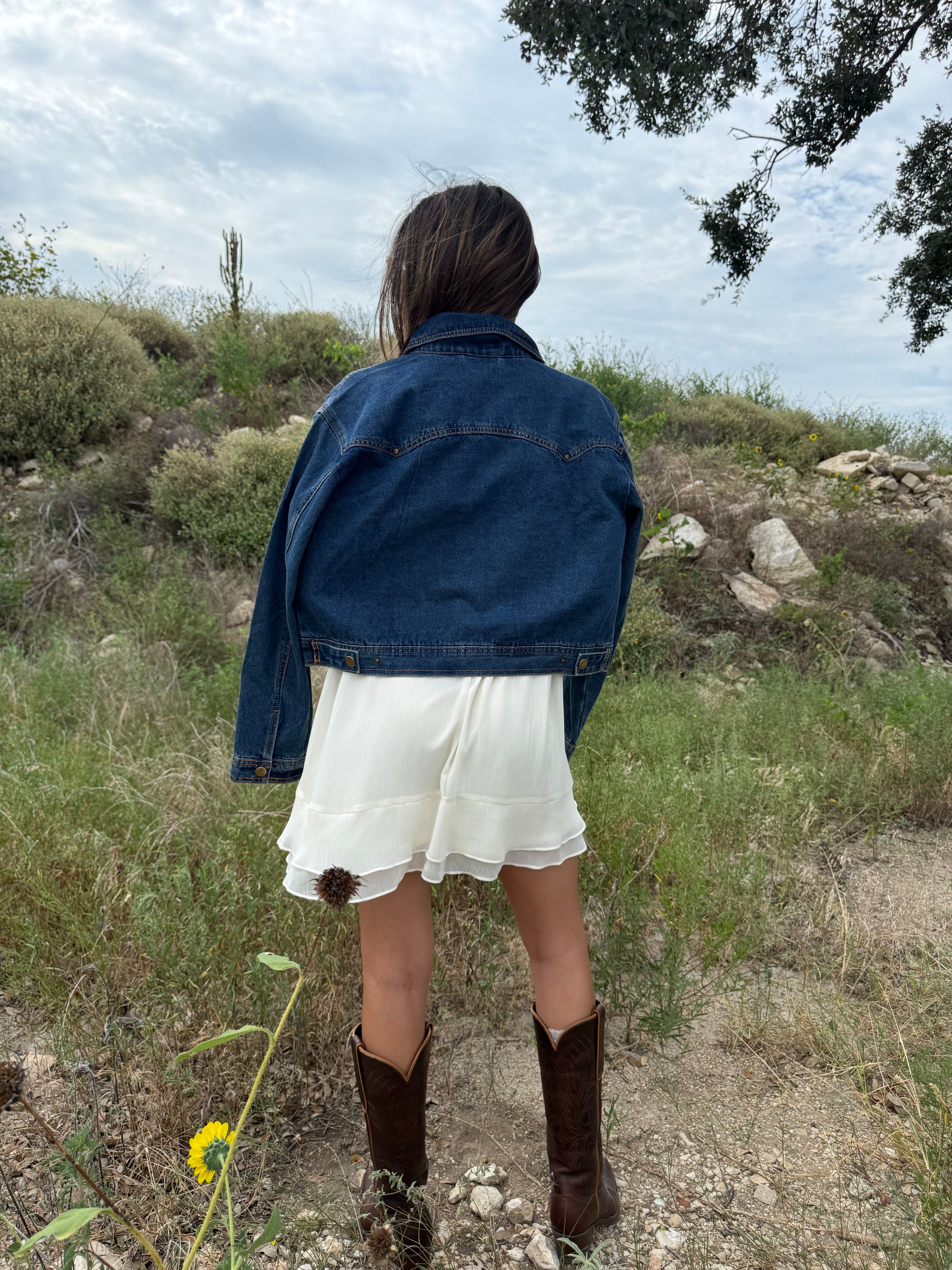 A woman viewed from behind wearing a cropped blue denim jacket, a white tiered skirt, and brown cowboy boots, standing outdoors on a grassy hillside under a cloudy sky. Fall rodeo western Texas style 