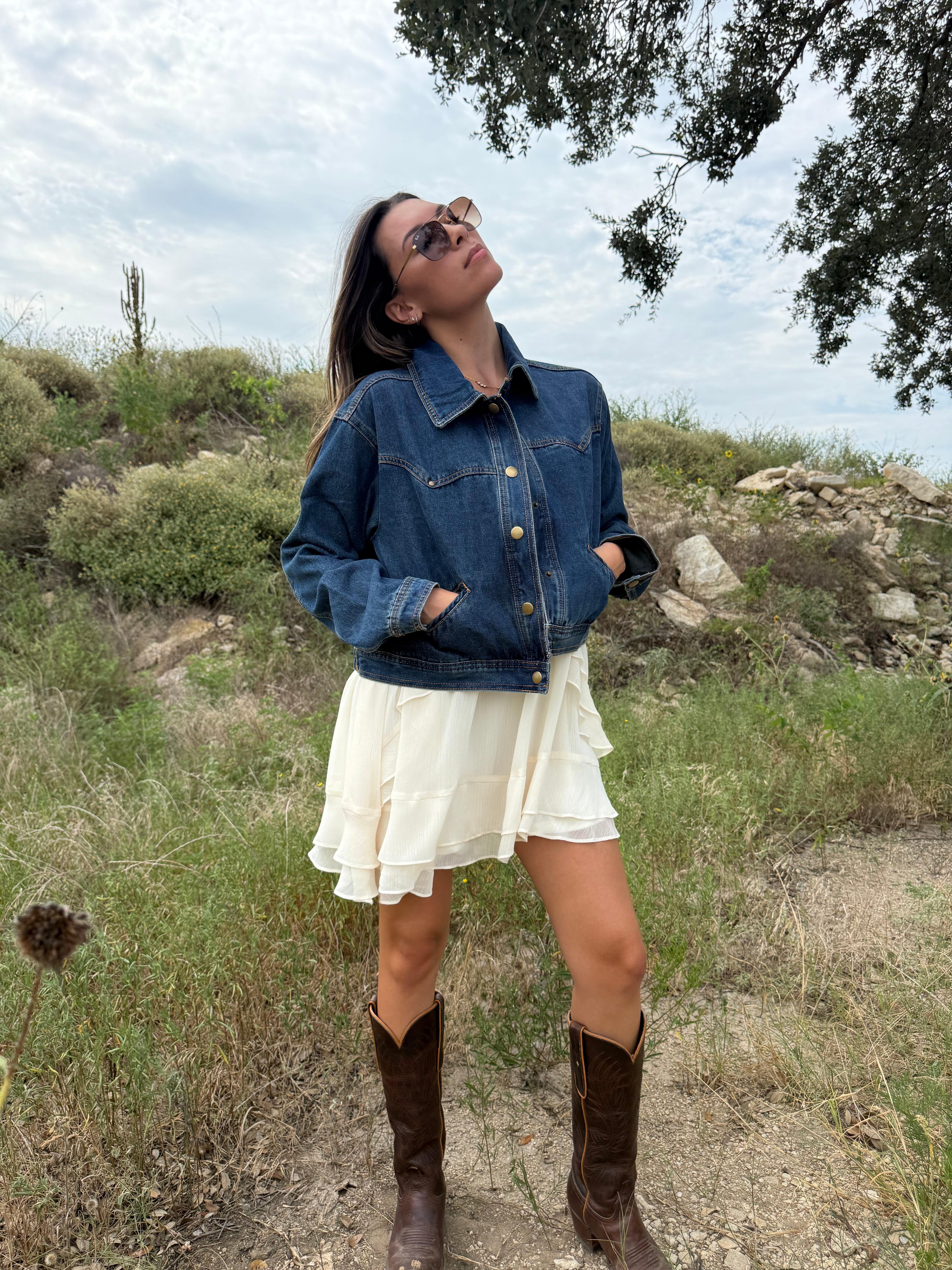 A woman wearing a dark blue denim jacket, cream ruffled mini dress, and brown cowboy boots stands outdoors in a grassy, rocky landscape under a cloudy sky. Fall Style Texas 