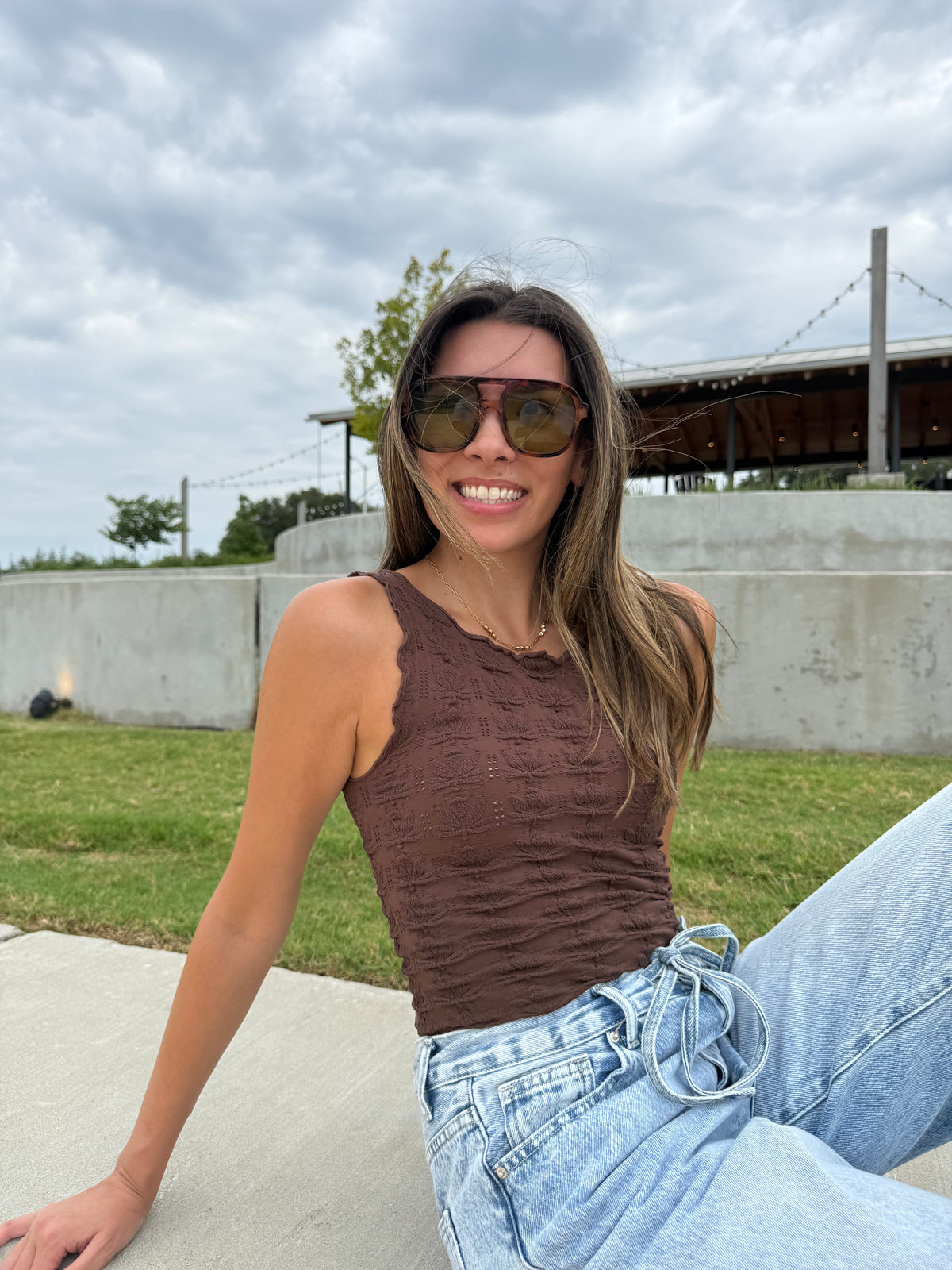 A woman wearing a brown textured sleeveless top and light blue jeans sits outdoors on a concrete surface under a cloudy sky.