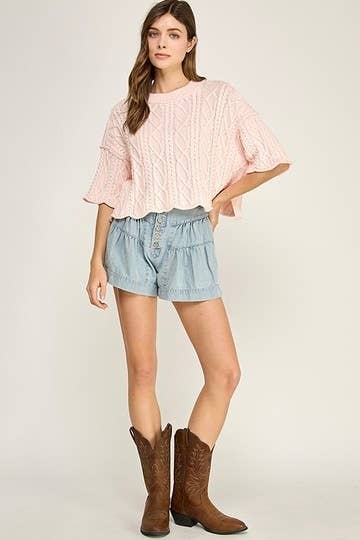 A woman modeling a light pink cable-knit short-sleeve top with ruffled edges, paired with light blue denim shorts and brown cowboy boots, against a plain light gray studio background.