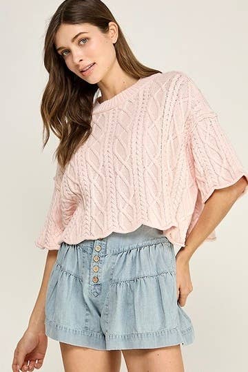 Woman wearing a pink cable knit top and light blue denim shorts on a plain background