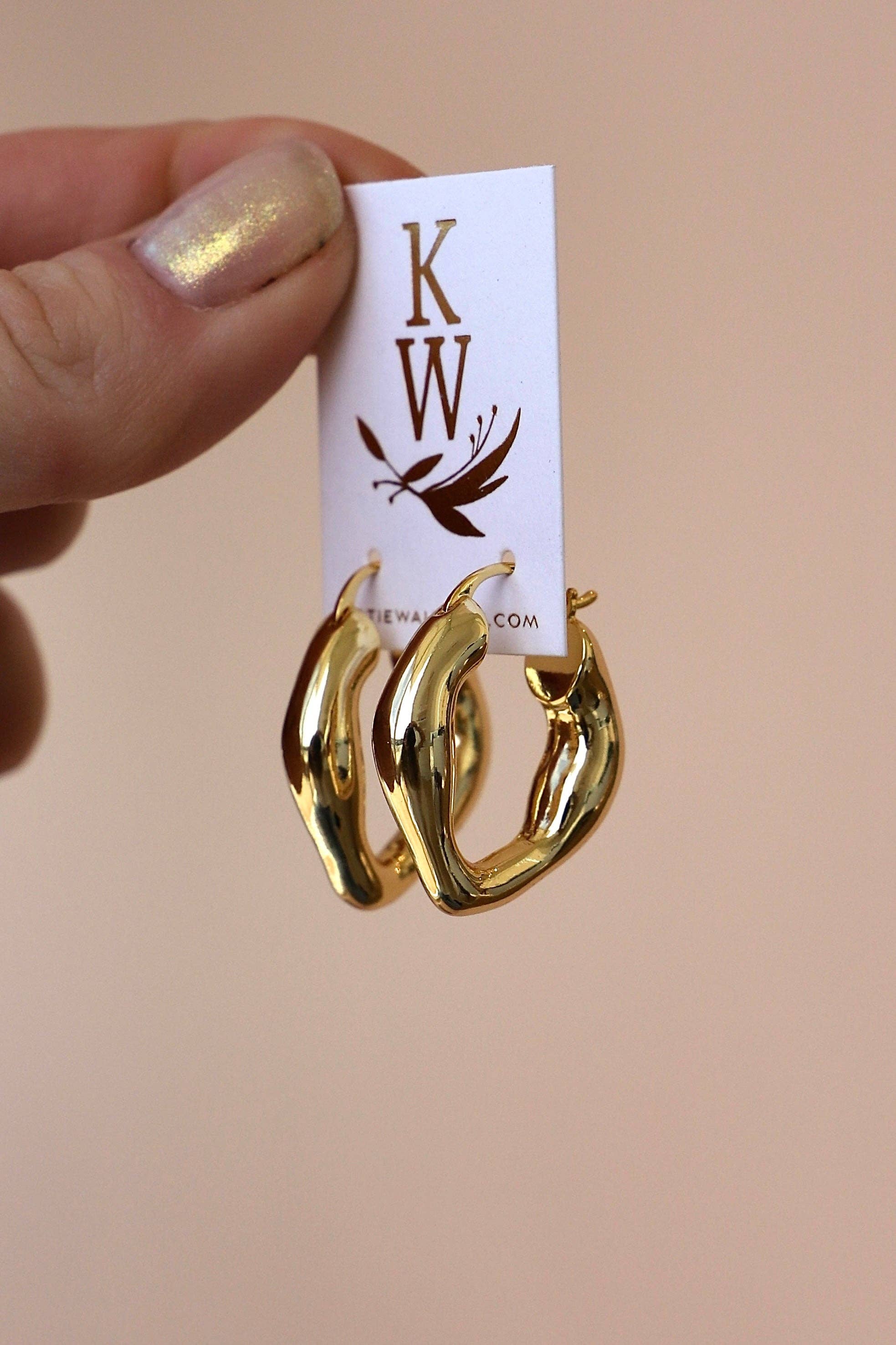 Gold chunky hoop earrings on a card with 'KW' branding held by a hand against a beige background