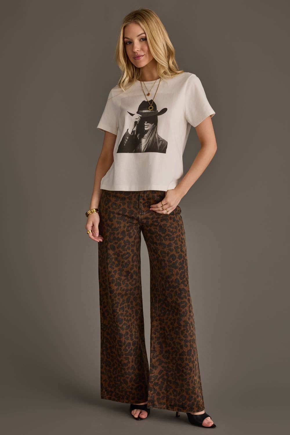 A woman modeling a white graphic t-shirt with a black-and-white portrait print and brown leopard-print wide-leg pants, standing against a solid dark gray studio background.