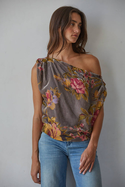 A woman wearing a gray off-the-shoulder top with a pink and yellow floral print, paired with blue jeans, standing against a plain light gray wall.