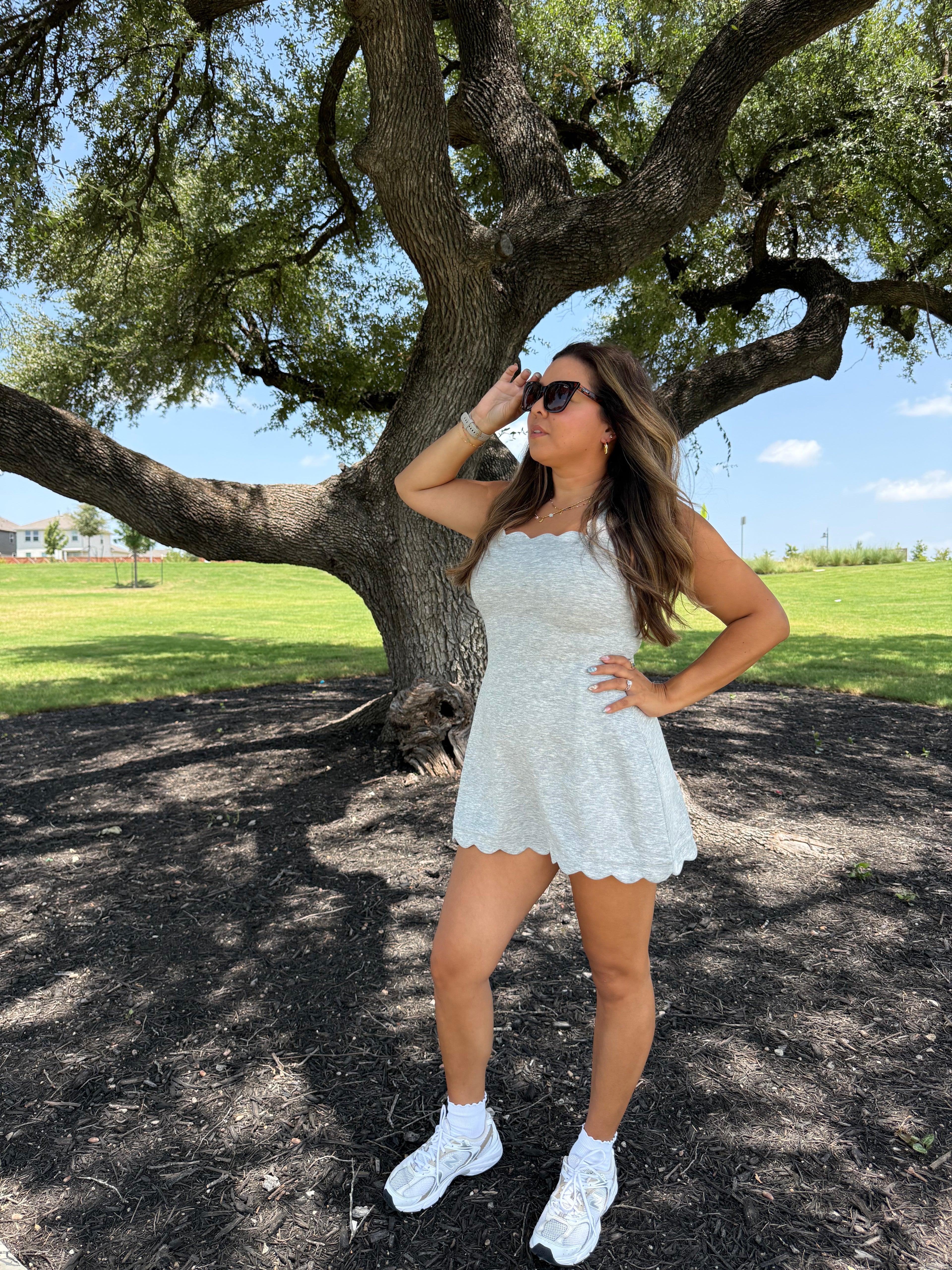 A woman posing outdoors under a large tree wearing a white lace mini dress, white sneakers, and white socks.