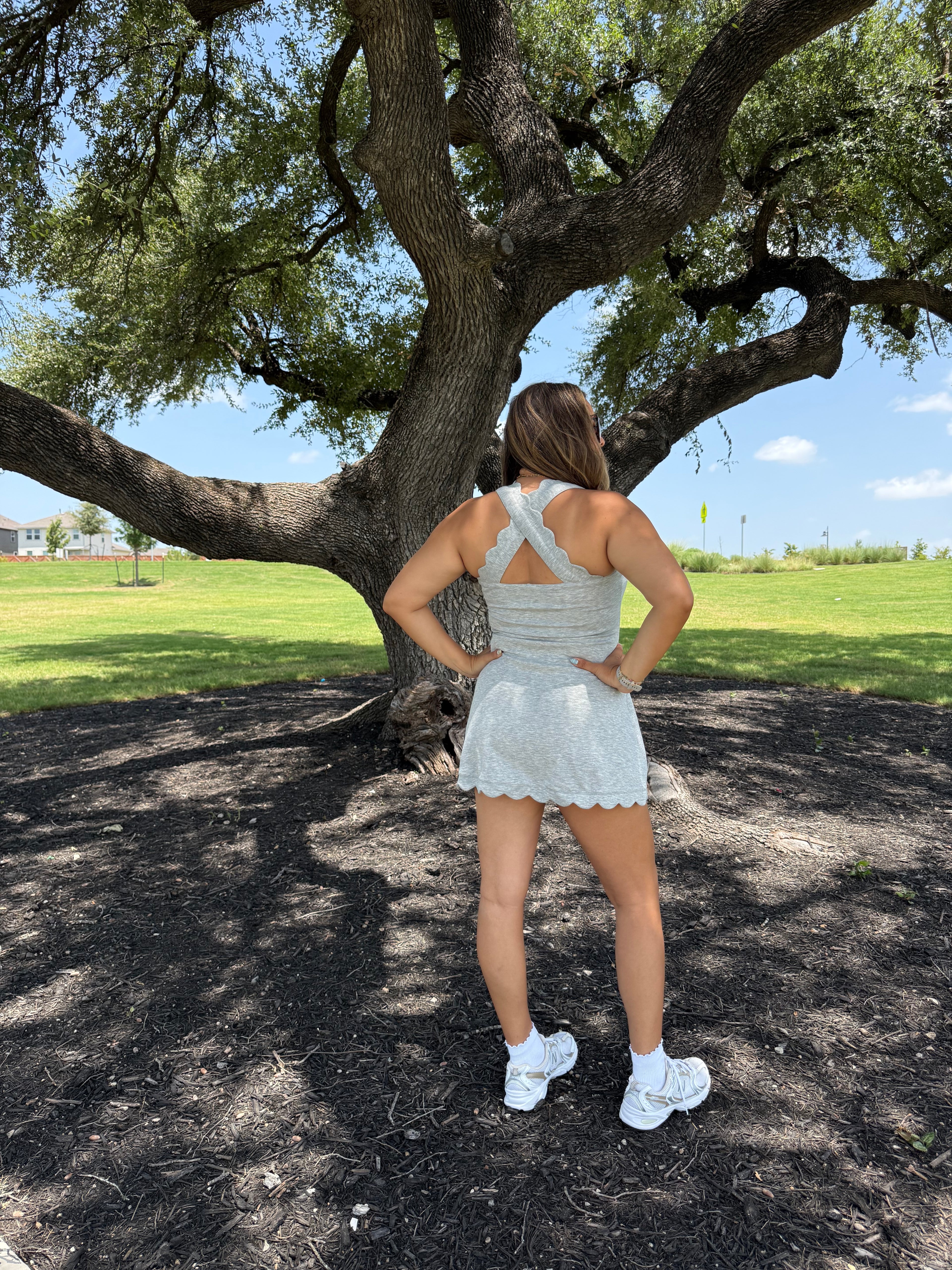 A woman stands with her back to the camera under a large tree in a park, wearing a white sleeveless athletic romper with a scalloped hem and white sneakers.