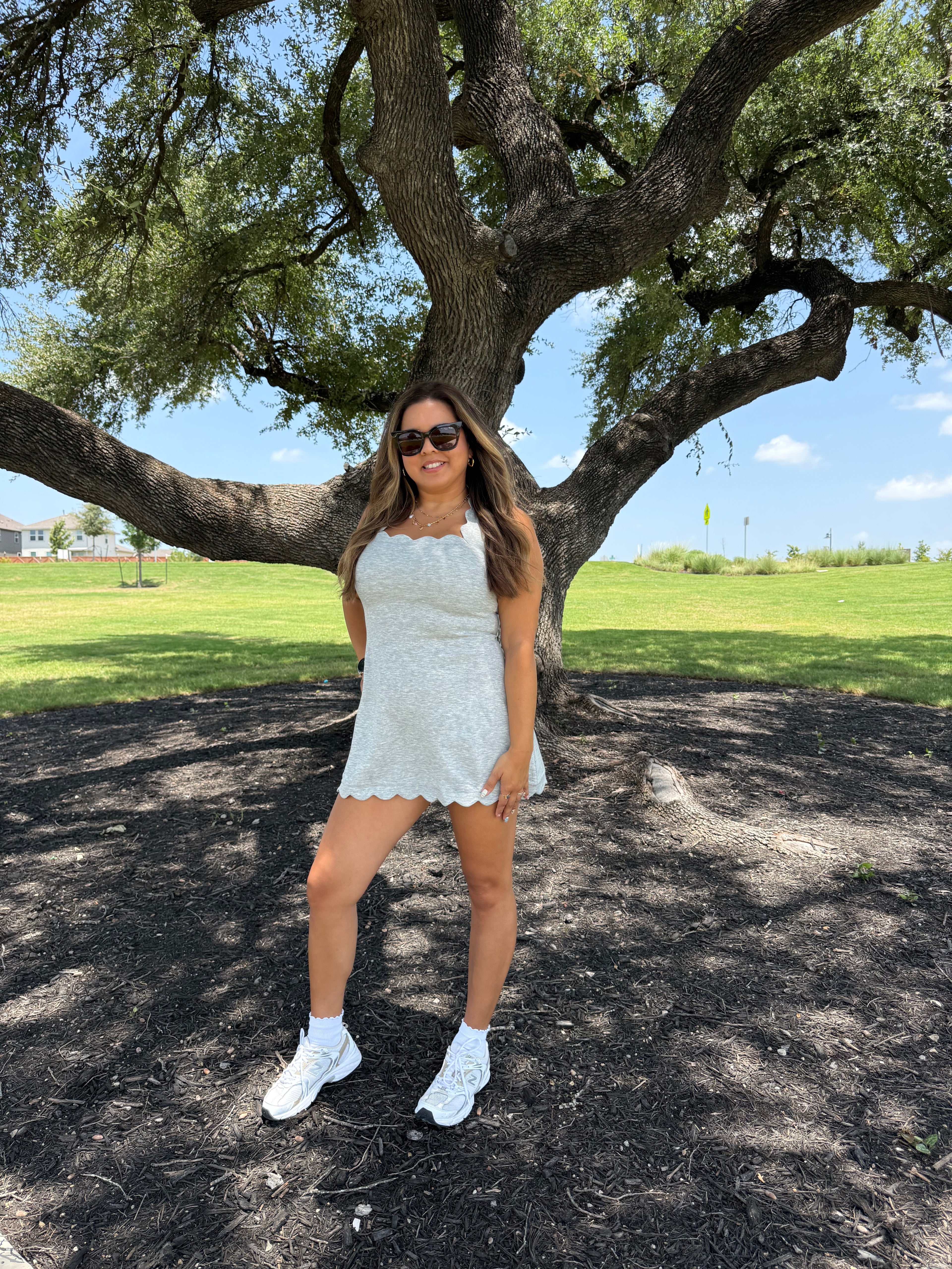 A woman wearing a scalloped grey lace athletic mini dress, white sneakers, and sunglasses stands under a large tree in a park with green grass and blue sky in the background.
