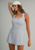 A woman modeling a light gray heather sleeveless mini dress with a scalloped neckline and hem, paired with an off-white baseball cap, against a solid light gray studio background. Athleisure