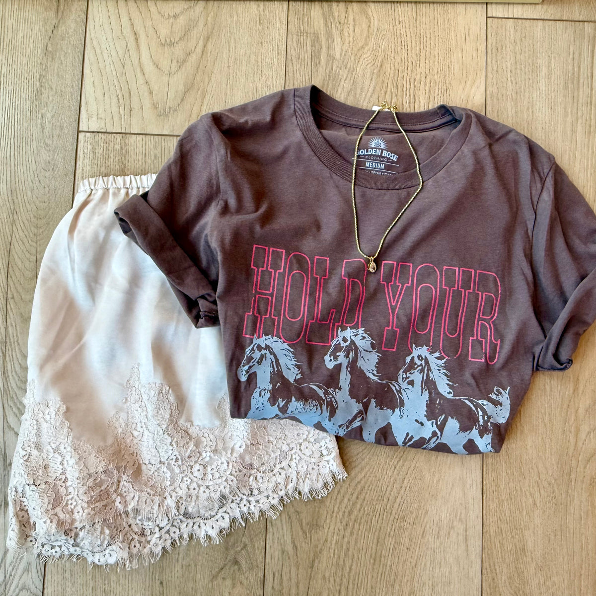 A brown short-sleeve graphic T-shirt with rolled sleeves, featuring the words 'HOLD YOUR' in pink outline above a gray horse silhouette print, styled flat on a light wood floor with a cream lace skirt and a gold necklace.