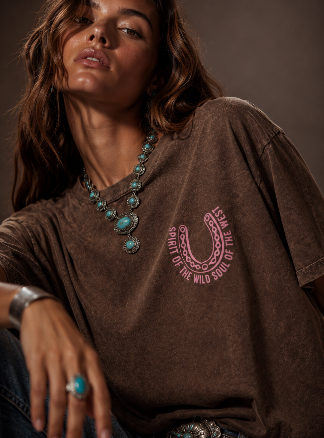Woman wearing a brown t-shirt with a horseshoe design and turquoise jewelry against a dark background