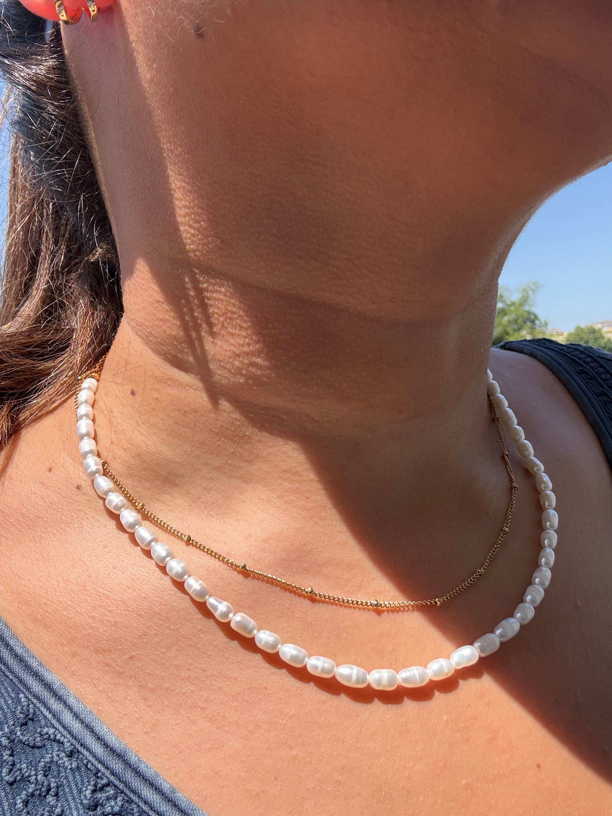 A close-up of a person's neck and upper chest wearing two delicate necklaces: a white pearl strand and a thin gold chain with small bead accents.