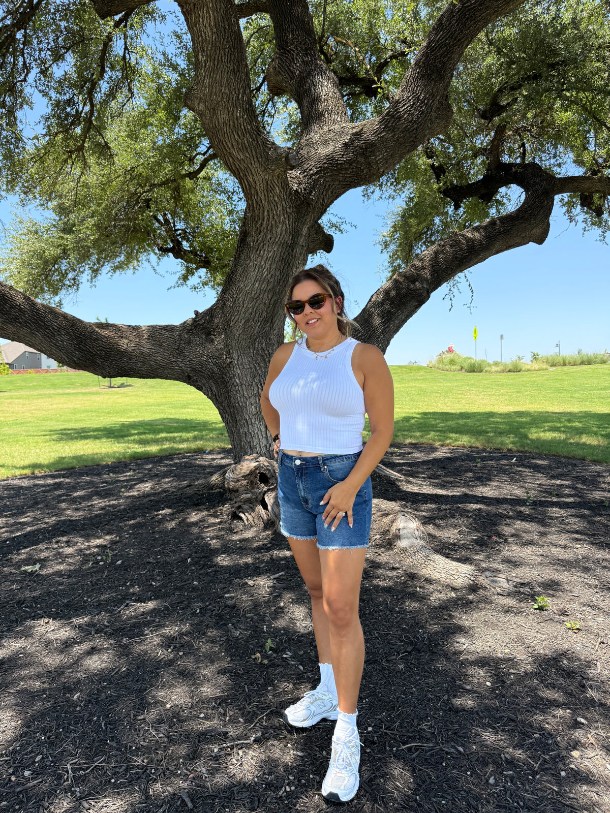 A woman wearing a white sleeveless top, denim shorts, white socks, and white sneakers poses outdoors under a large tree on a dirt path with green grass in the background.