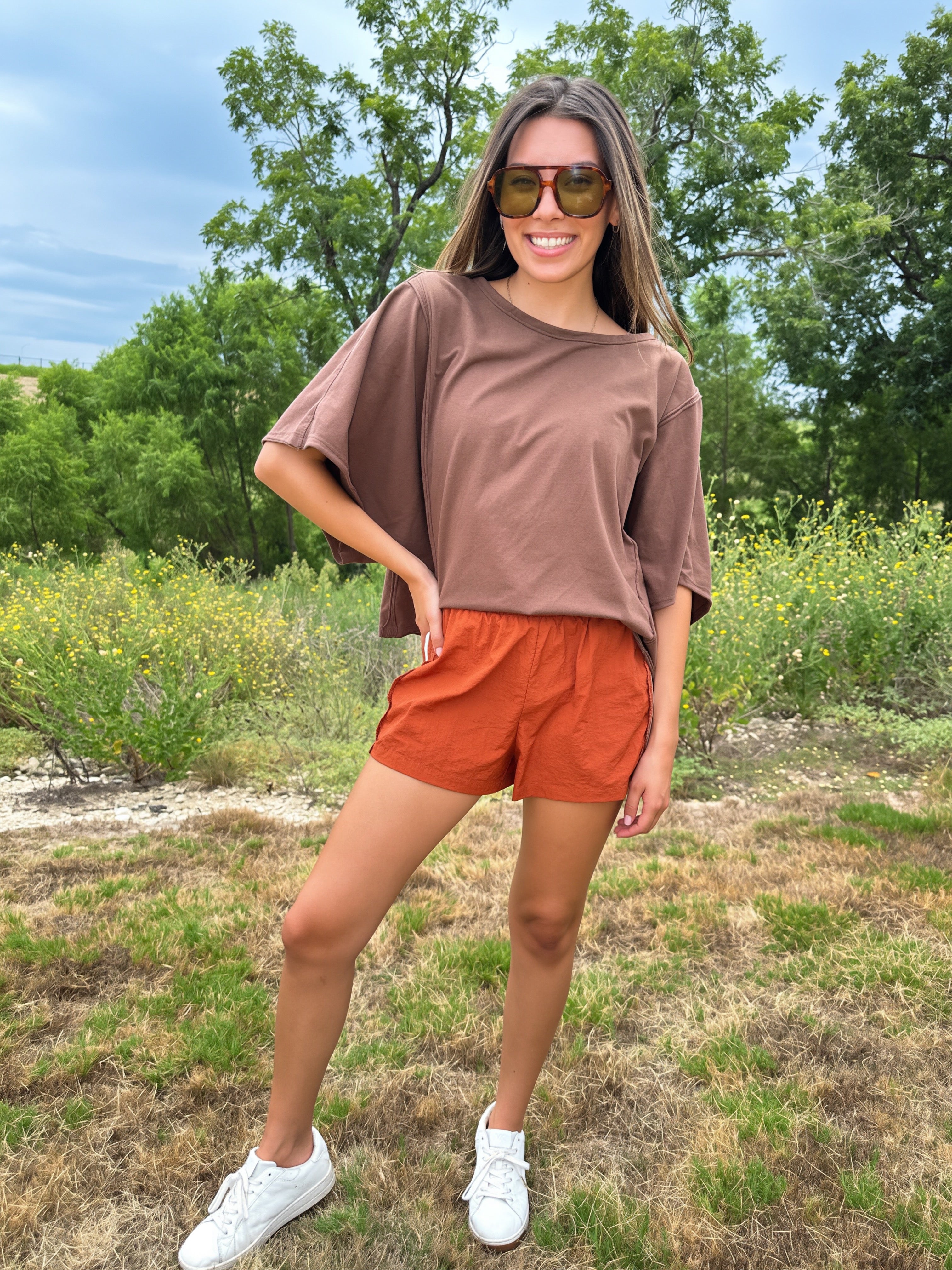 A woman wearing a brown short-sleeve t-shirt and rust-orange shorts stands outdoors in a grassy field with trees in the background, wearing white sneakers and sunglasses.