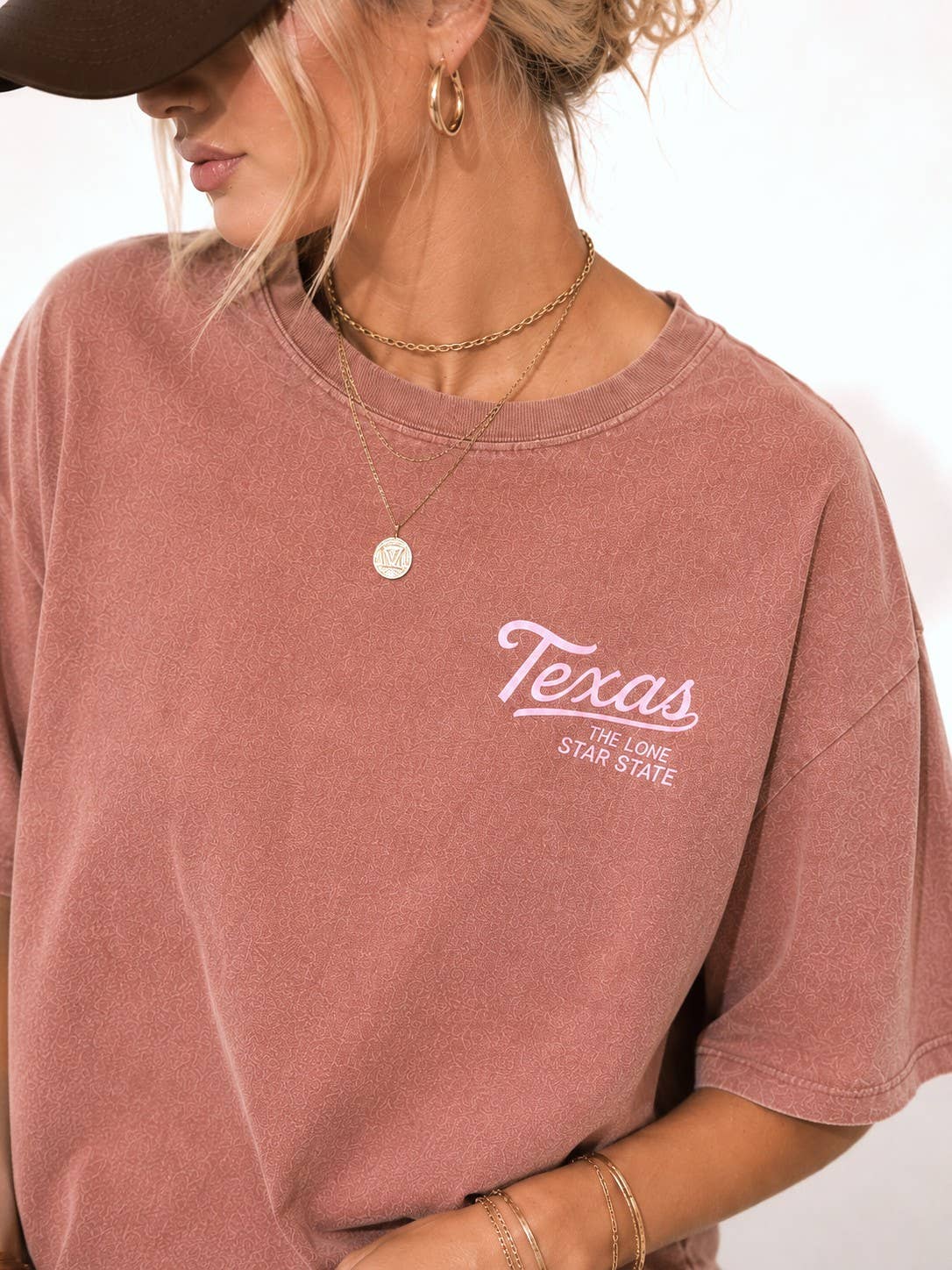 A woman wearing a faded dusty mauve rose short sleeve t-shirt with 'Texas' printed in white script on the left chest, styled with layered gold necklaces and a dark brown baseball cap.