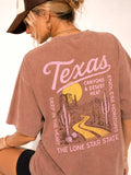 A woman wearing a faded mauve pink oversized graphic T-shirt, shown from the back. The shirt features a desert landscape print with cacti, a winding yellow road, a yellow sun, and pink text reading 'Texas' and 'Canyons & Desert Heat'.