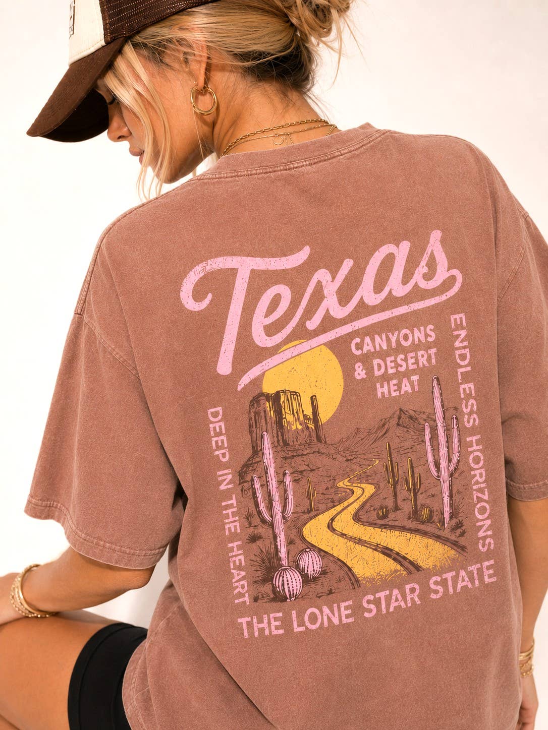 A woman wearing a faded mauve pink oversized graphic T-shirt, shown from the back. The shirt features a desert landscape print with cacti, a winding yellow road, a yellow sun, and pink text reading 'Texas' and 'Canyons & Desert Heat'.