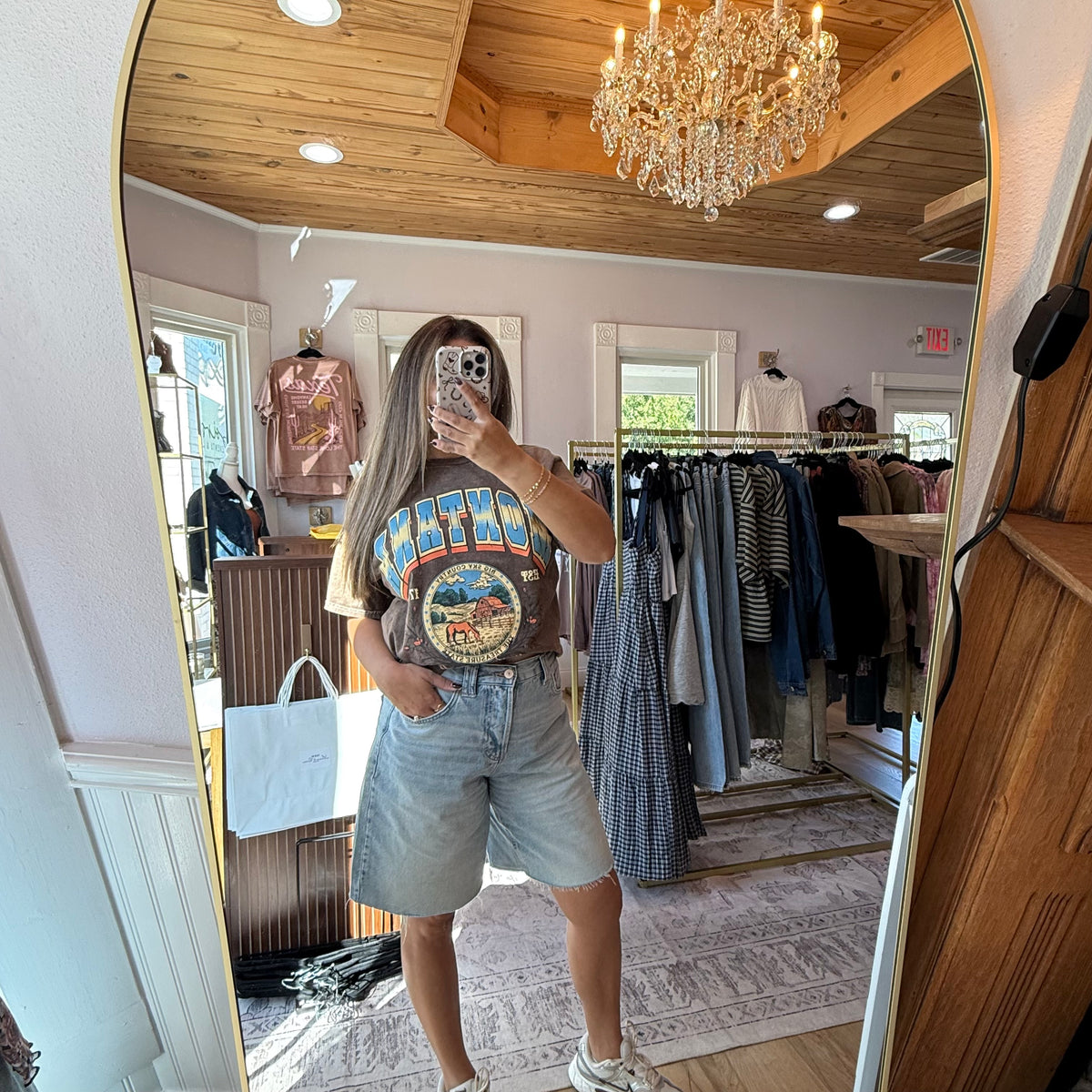 A woman takes a mirror selfie in a boutique while wearing a brown Montana graphic t-shirt and light blue denim shorts. The boutique interior features a wooden ceiling, a crystal chandelier, and clothing racks in the background.