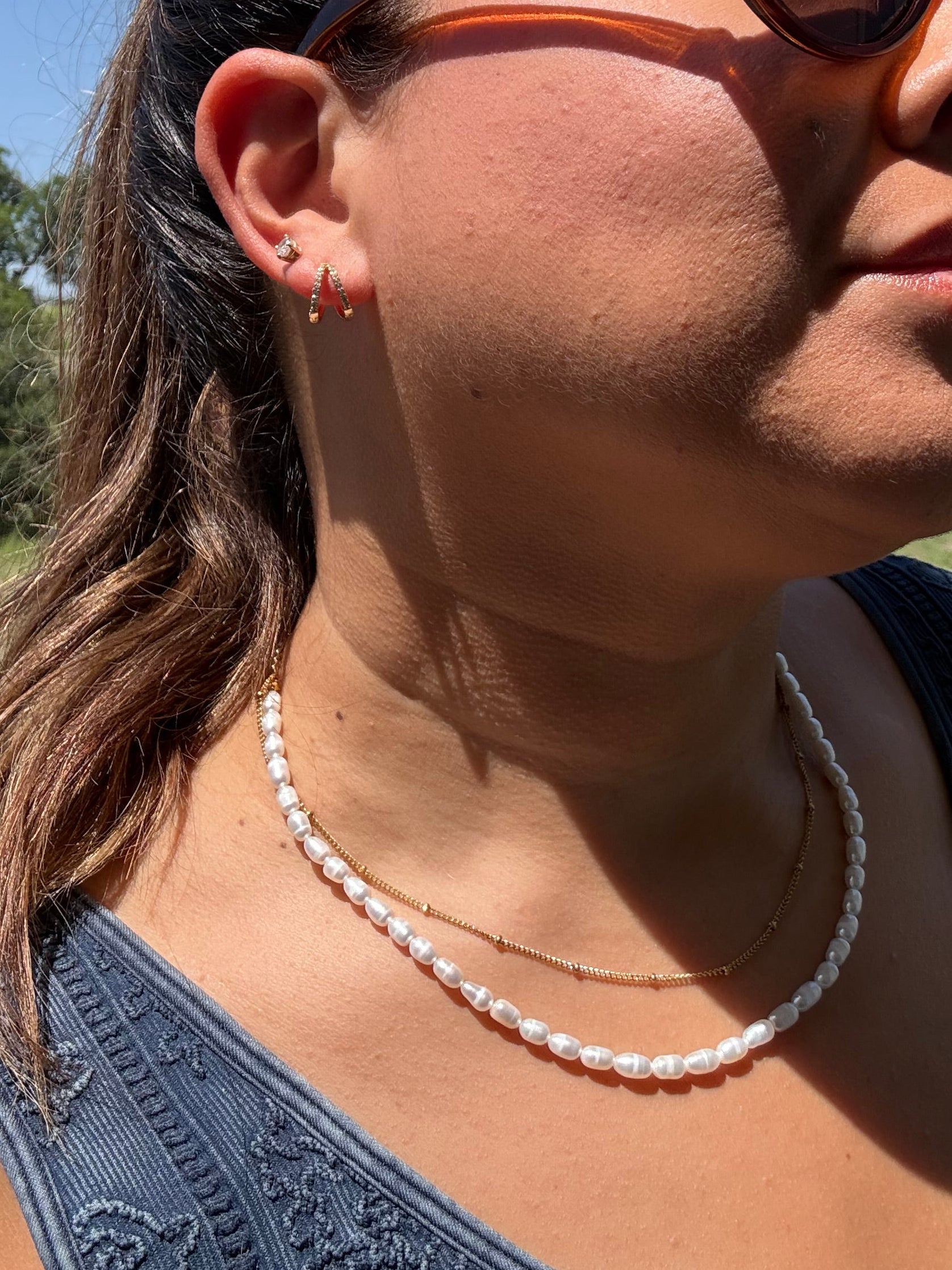 A close-up outdoor shot of a woman wearing brown cat-eye sunglasses, a white pearl necklace, and a blue lace-trim top against a blue sky and green field background.