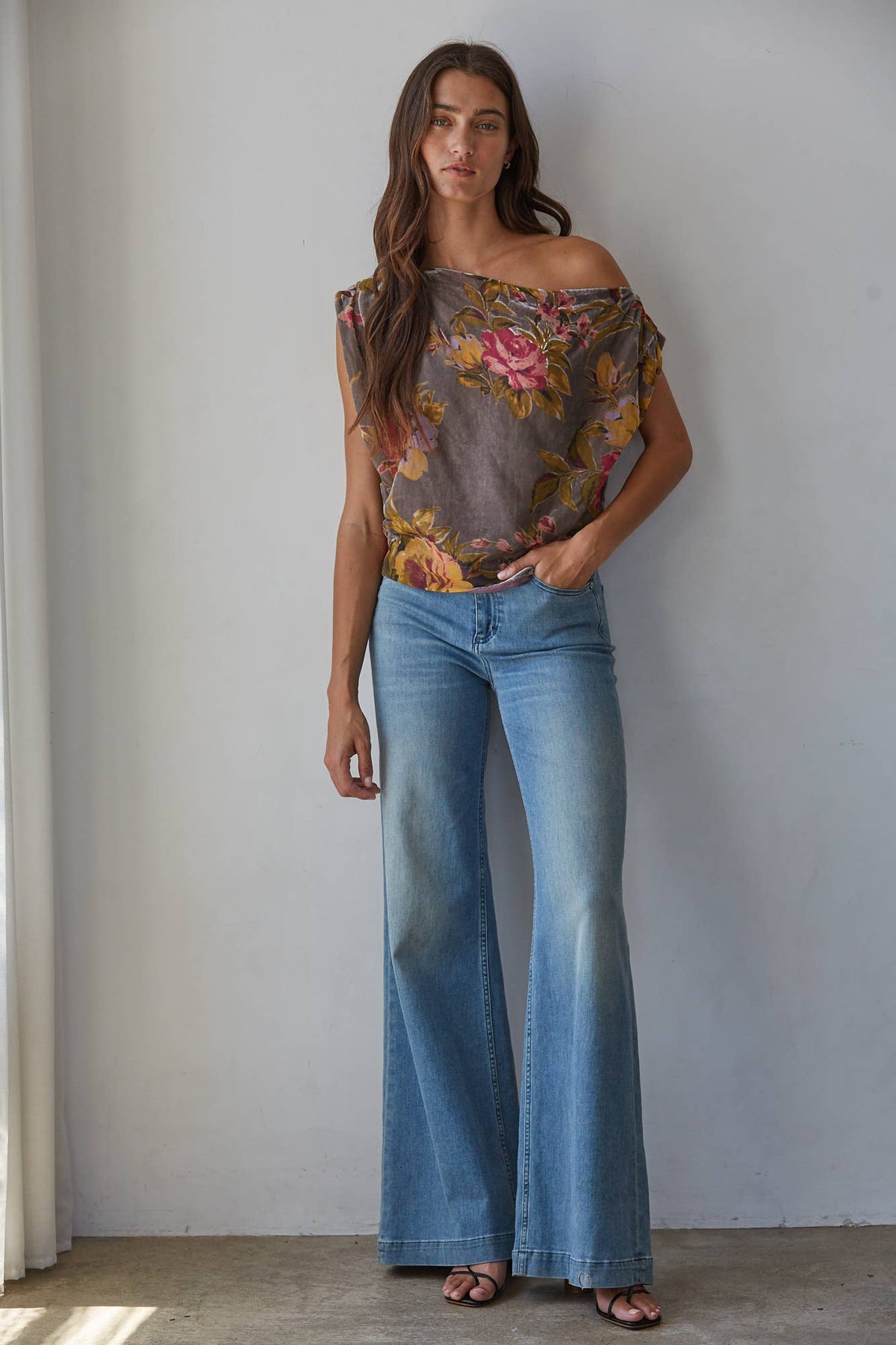 A woman stands against a light gray wall wearing a sheer off-the-shoulder floral top with blue wide-leg jeans and brown sandals.