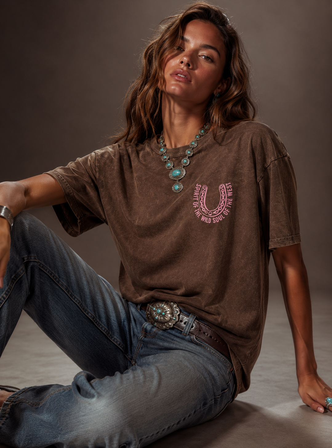 Person wearing a brown t-shirt with a graphic design and blue jeans, sitting on the floor against a neutral background.
