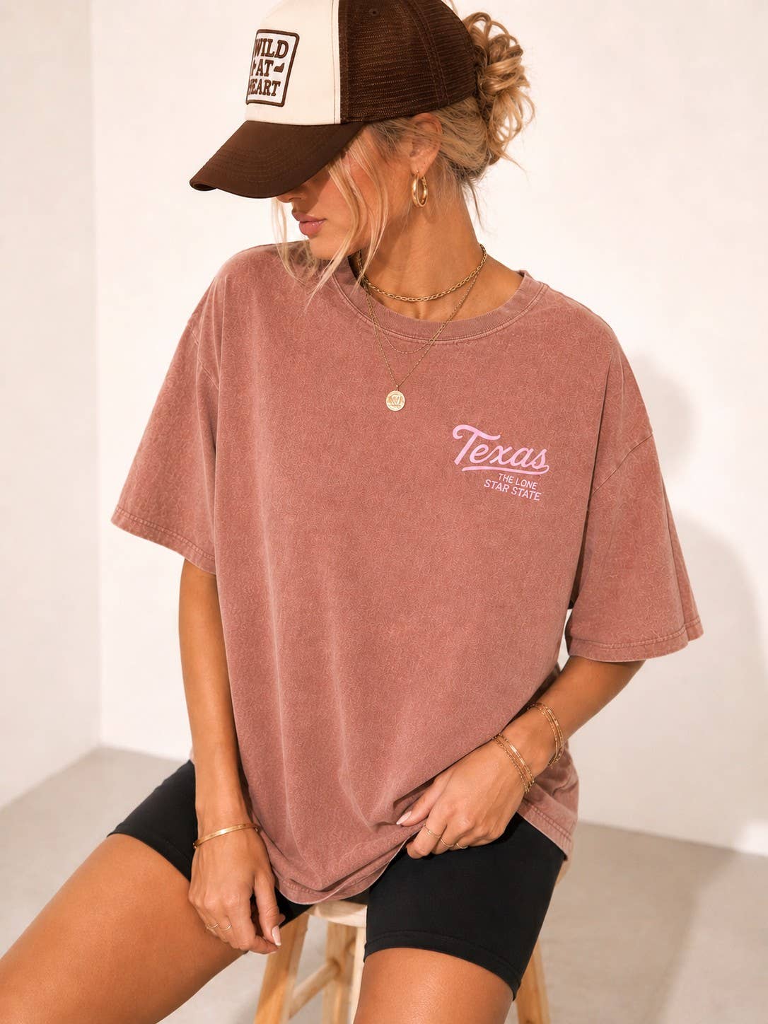 A woman wearing an oversized dusty rose short-sleeve t-shirt with a 'Texas Star Love' graphic on the chest, paired with black shorts and a brown and white trucker hat.