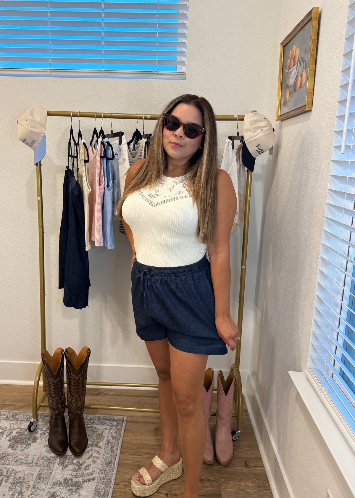 A woman modeling a Pale butter Yellow ribbed sleeveless top with a lace-up neckline and dark blue denim shorts, standing in front of a gold clothing rack in a room with windows and boots.