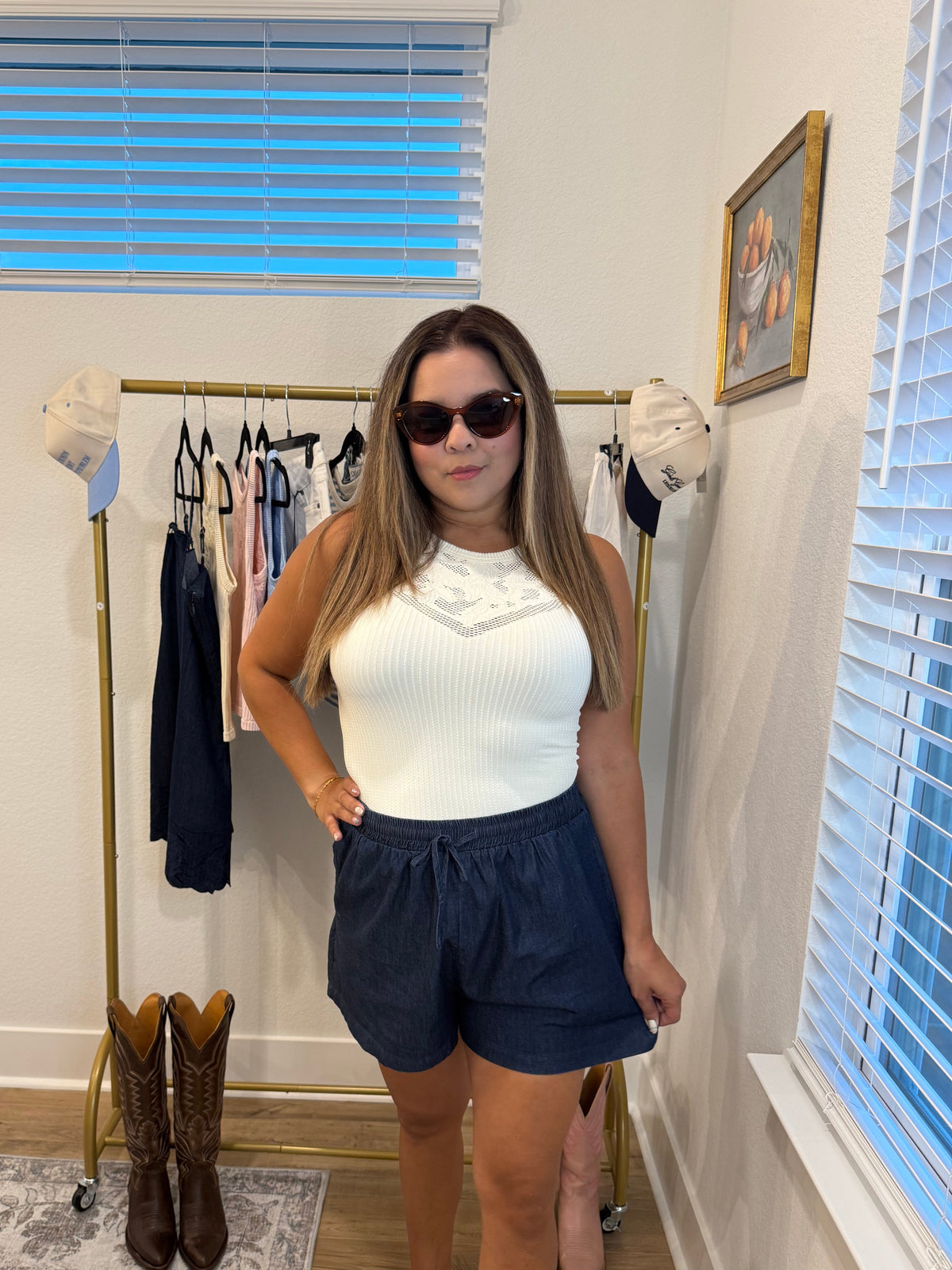 A woman wearing a pale butter Yellow ribbed tank top with a lace detail at the neckline and dark blue denim shorts, standing in front of a clothing rack in a home setting.