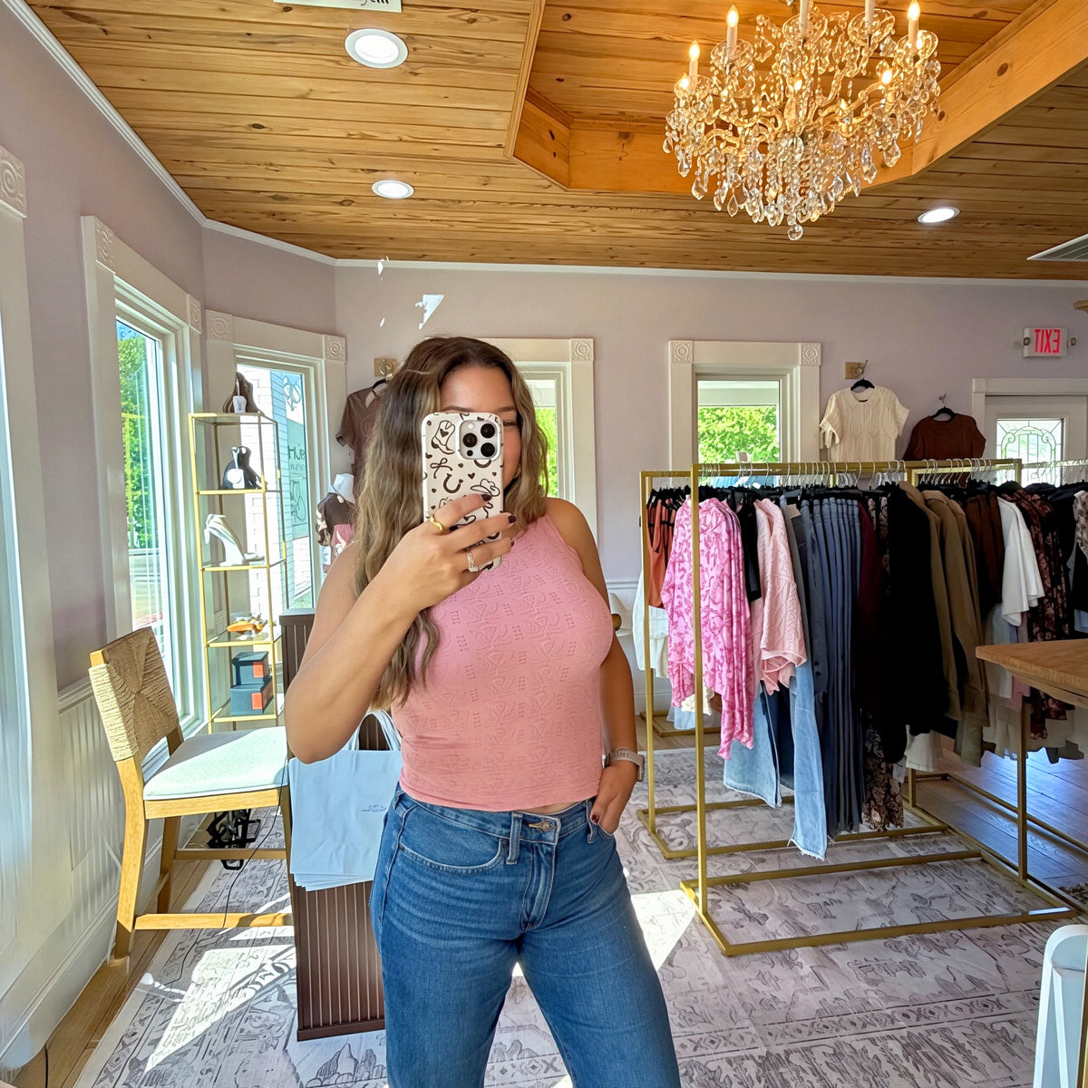 A woman takes a mirror selfie in a boutique, wearing a light pink ribbed tank top and blue jeans. The shop features a wooden ceiling, a large crystal chandelier, and clothing racks filled with garments.