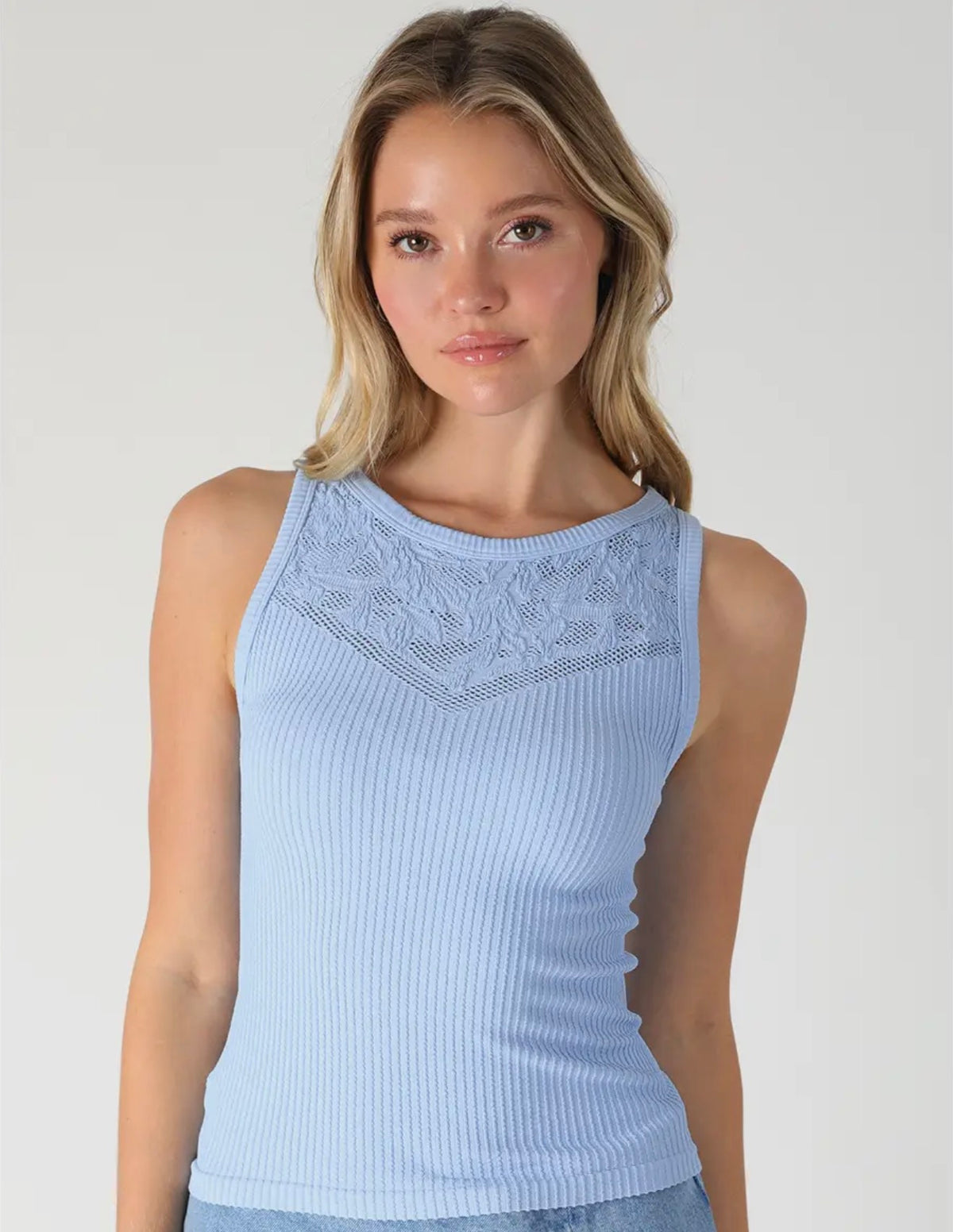 A woman wearing a light blue ribbed sleeveless top with a lace-like yoke detail, photographed against a plain light gray studio background.