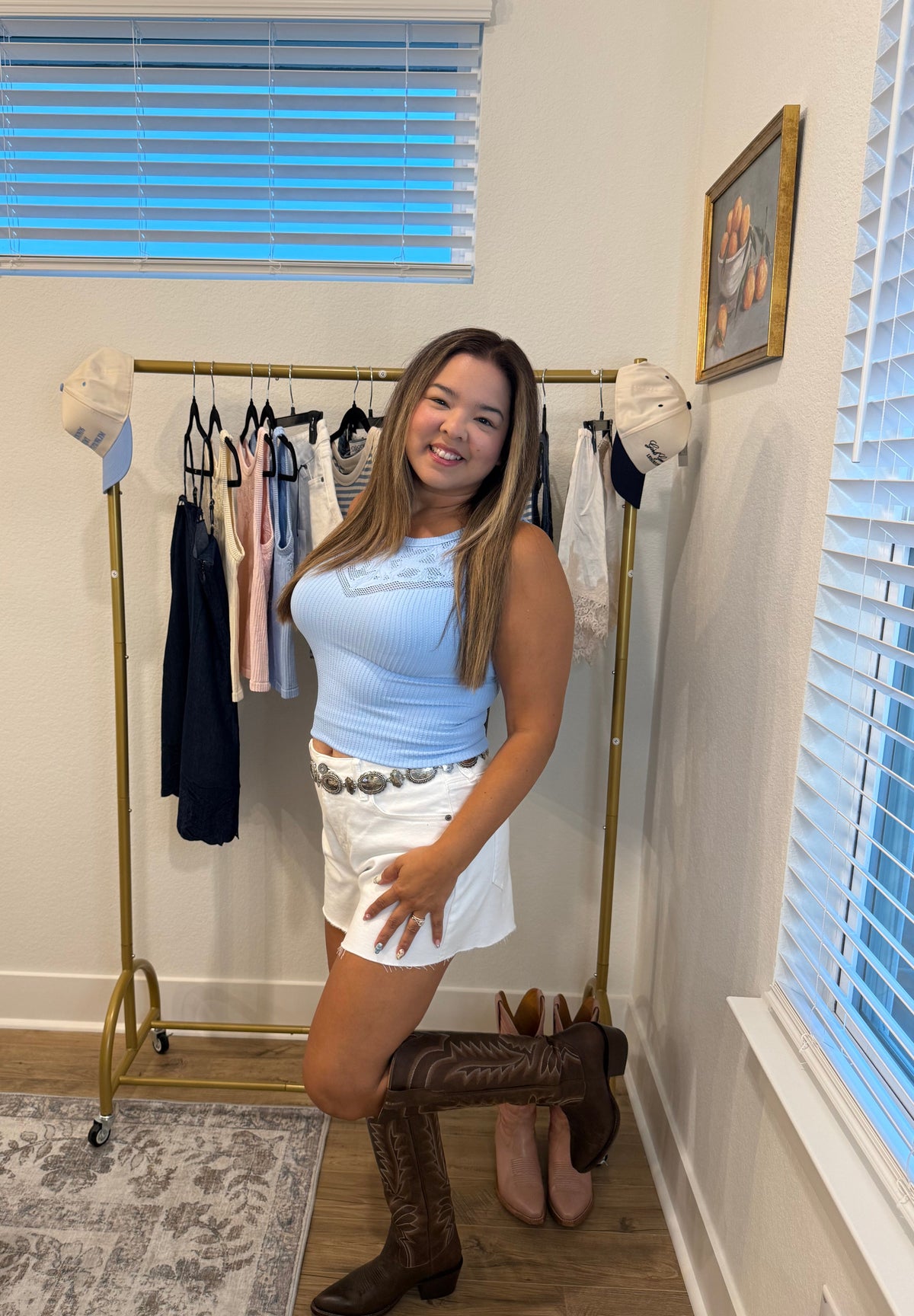 A woman posing in a white sleeveless top and white shorts with a black belt, paired with brown cowboy boots, standing in a room with a clothing rack and window blinds.