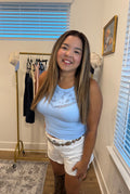 A woman wearing a light blue ribbed tank top with a white graphic print and white shorts, standing in a room with window blinds and a clothing rack.